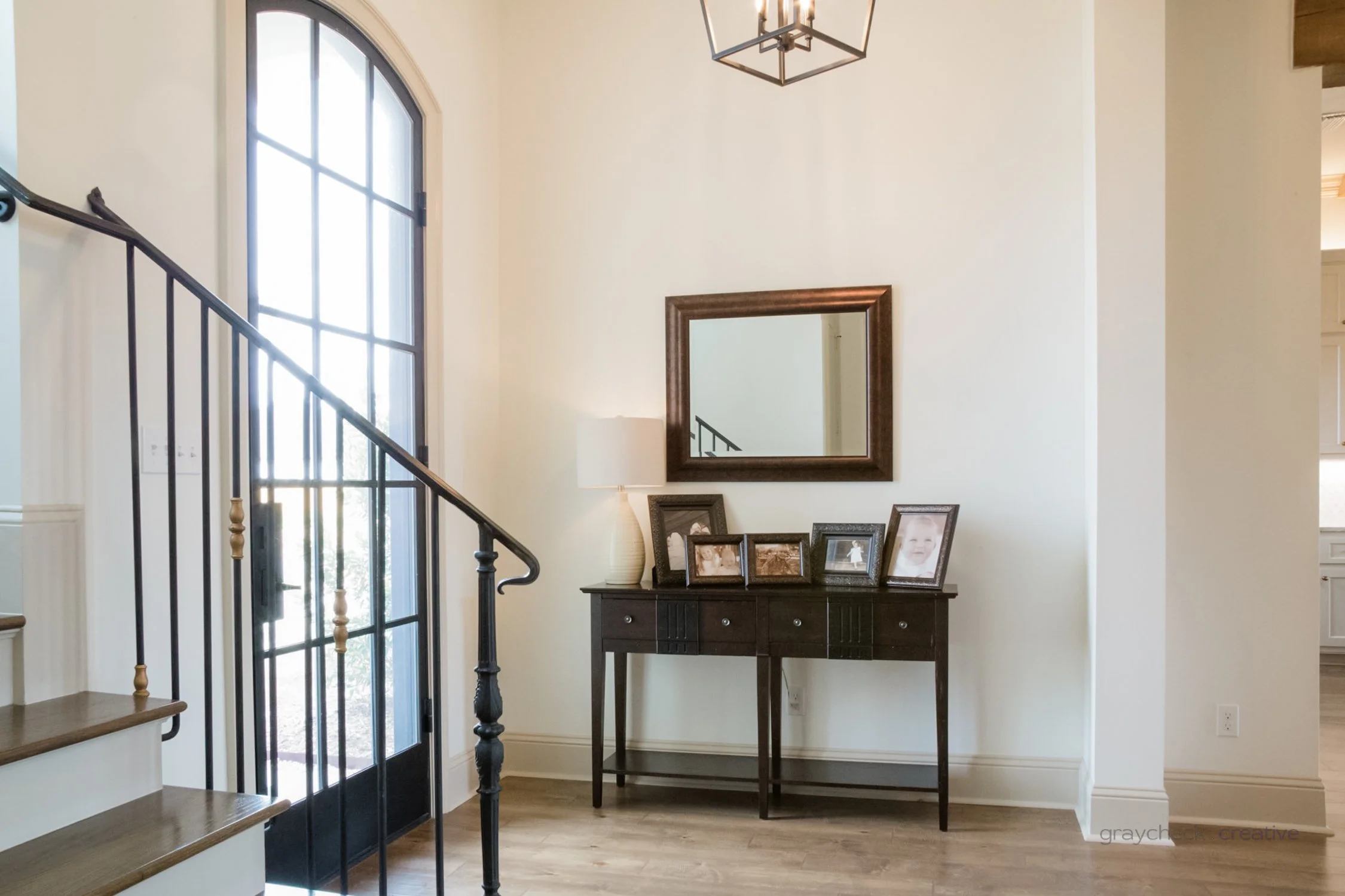 Entryway with a black console table holding framed photos, a lamp, and a mirror on the wall above, near a staircase and a large, arched window.