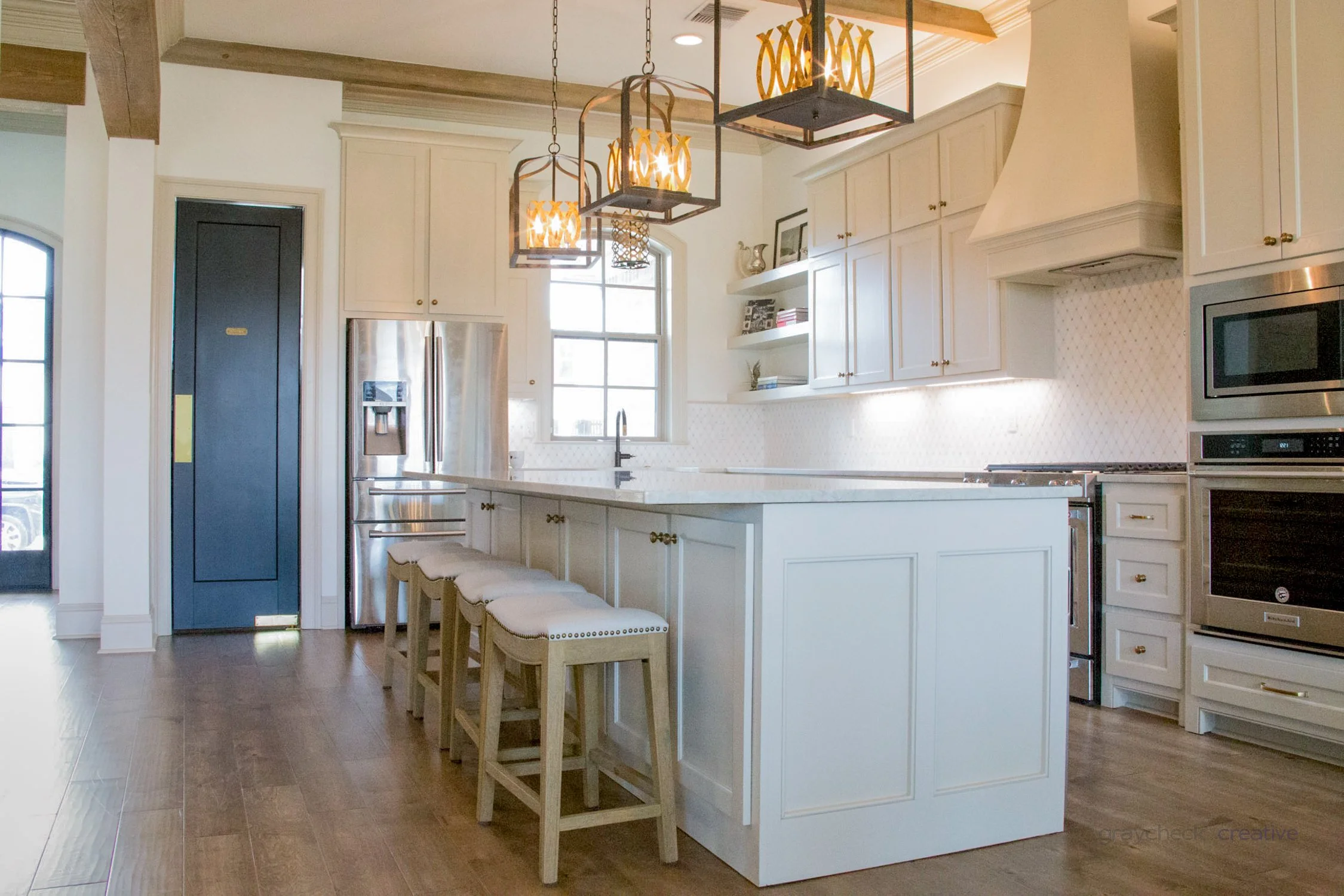 Modern kitchen with white cabinets, stainless steel refrigerator and appliances, a kitchen island with four bar stools, wooden flooring, and pendant lighting fixtures.