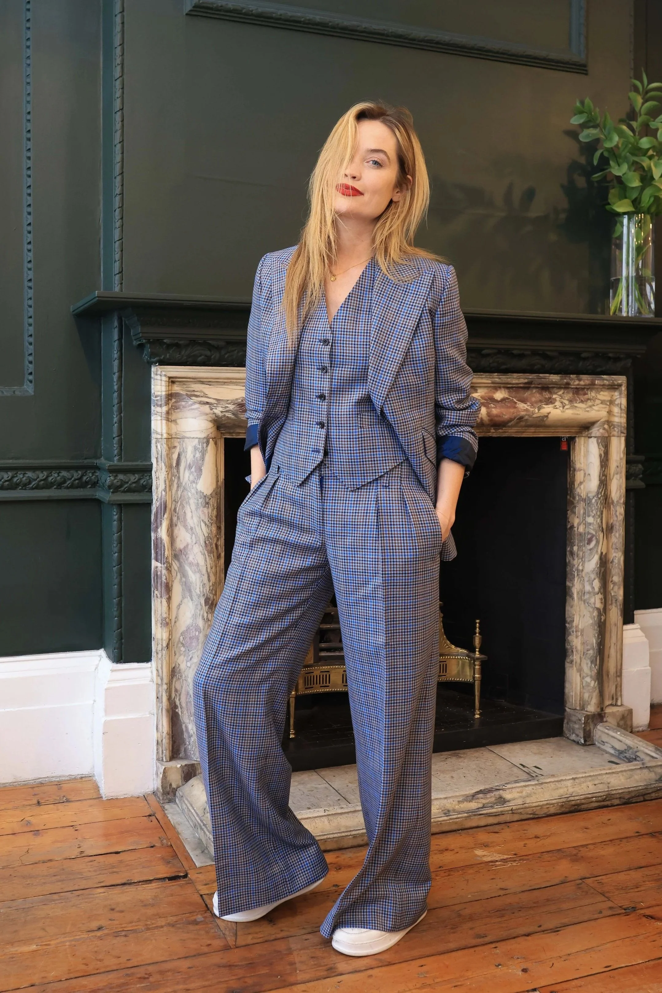 A woman with blonde hair wearing a plaid suit with wide-legged pants and a blazer, standing in front of a marble fire place with green walls and a plant on a table.
