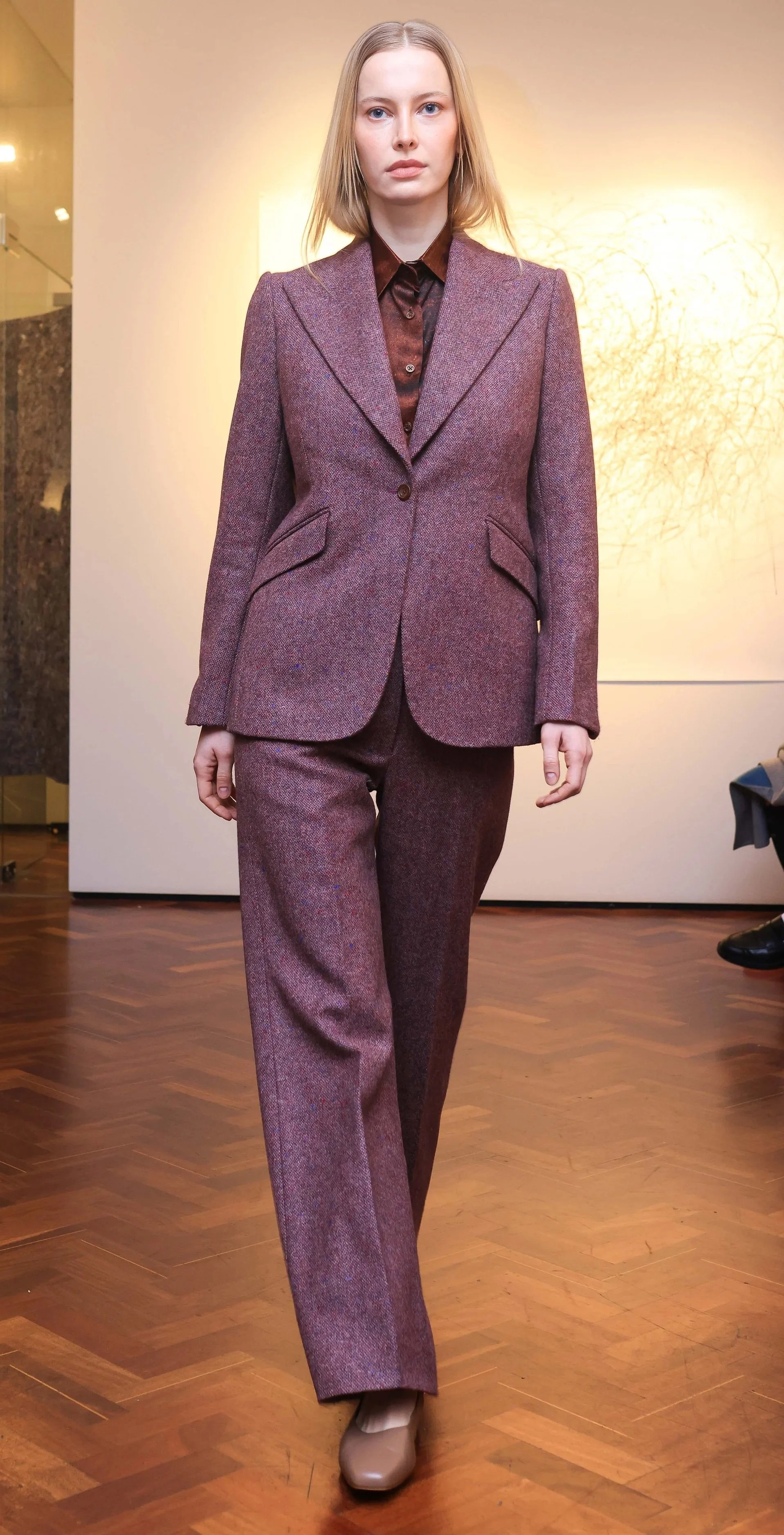 A woman walking on a wooden floor in a burgundy-colored suit with a blazer and matching pants.