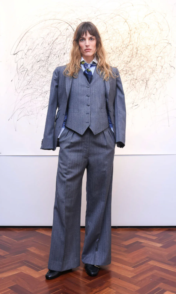 A woman standing in front of a wall with abstract scribbles, wearing a gray business suit with a vest, shirt, tie, and black shoes.