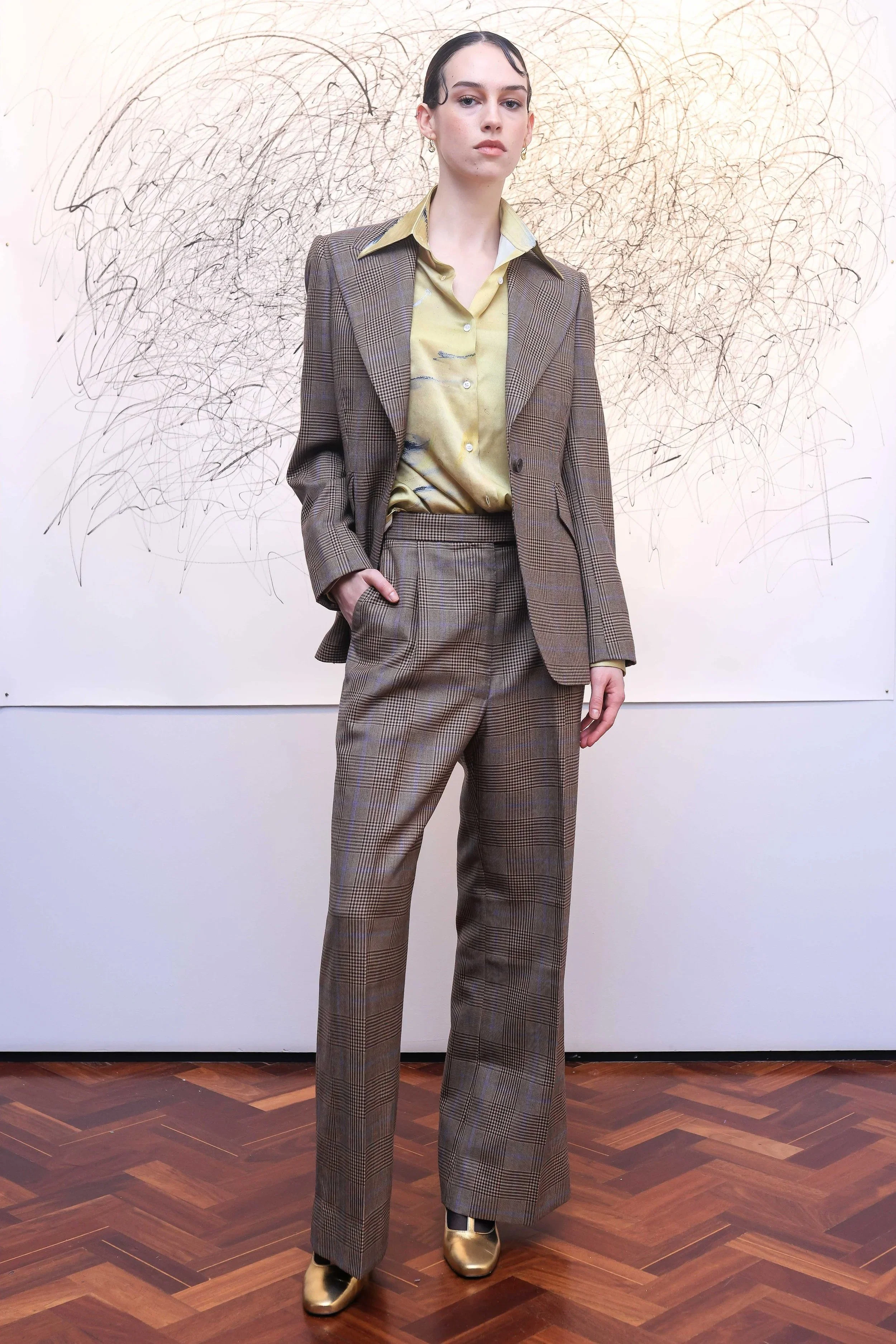 Woman dressed in a patterned plaid suit with a light yellow satin shirt, standing in front of a wall with abstract black scribbles, on a wooden floor.