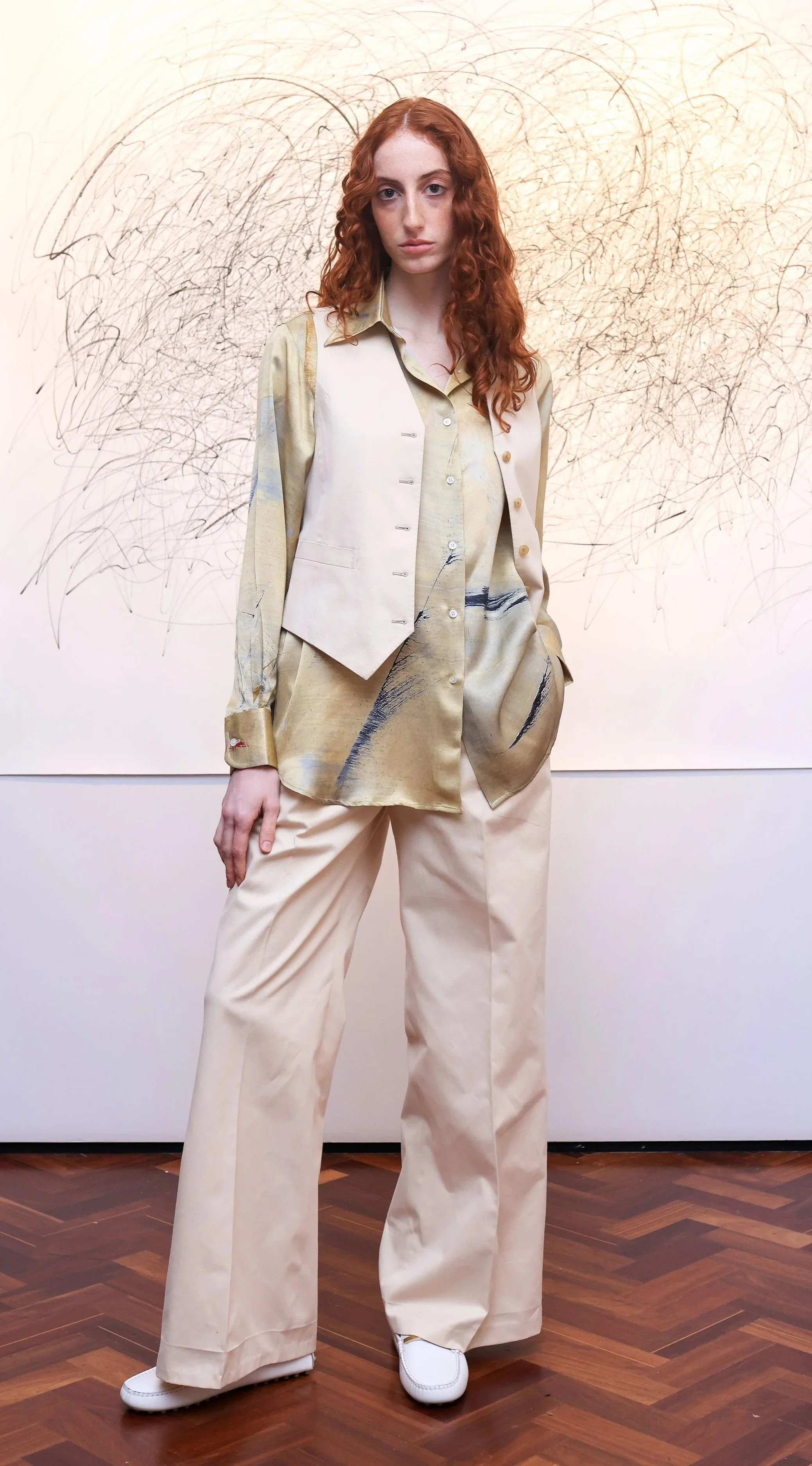 A young woman with long, curly red hair standing in front of an abstract line art background. She is wearing a beige vest, a patterned beige shirt, wide-leg beige trousers, and white shoes.