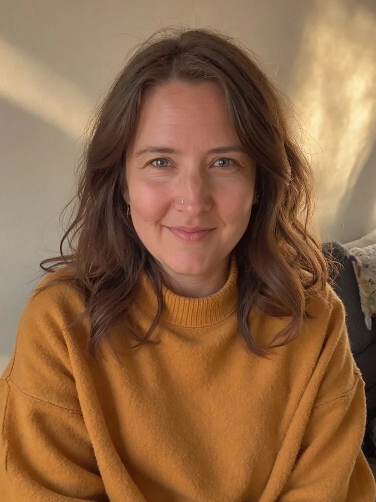 A picture of Ashley wearing a mustard-colored sweater, smiling indoors with warm lighting.