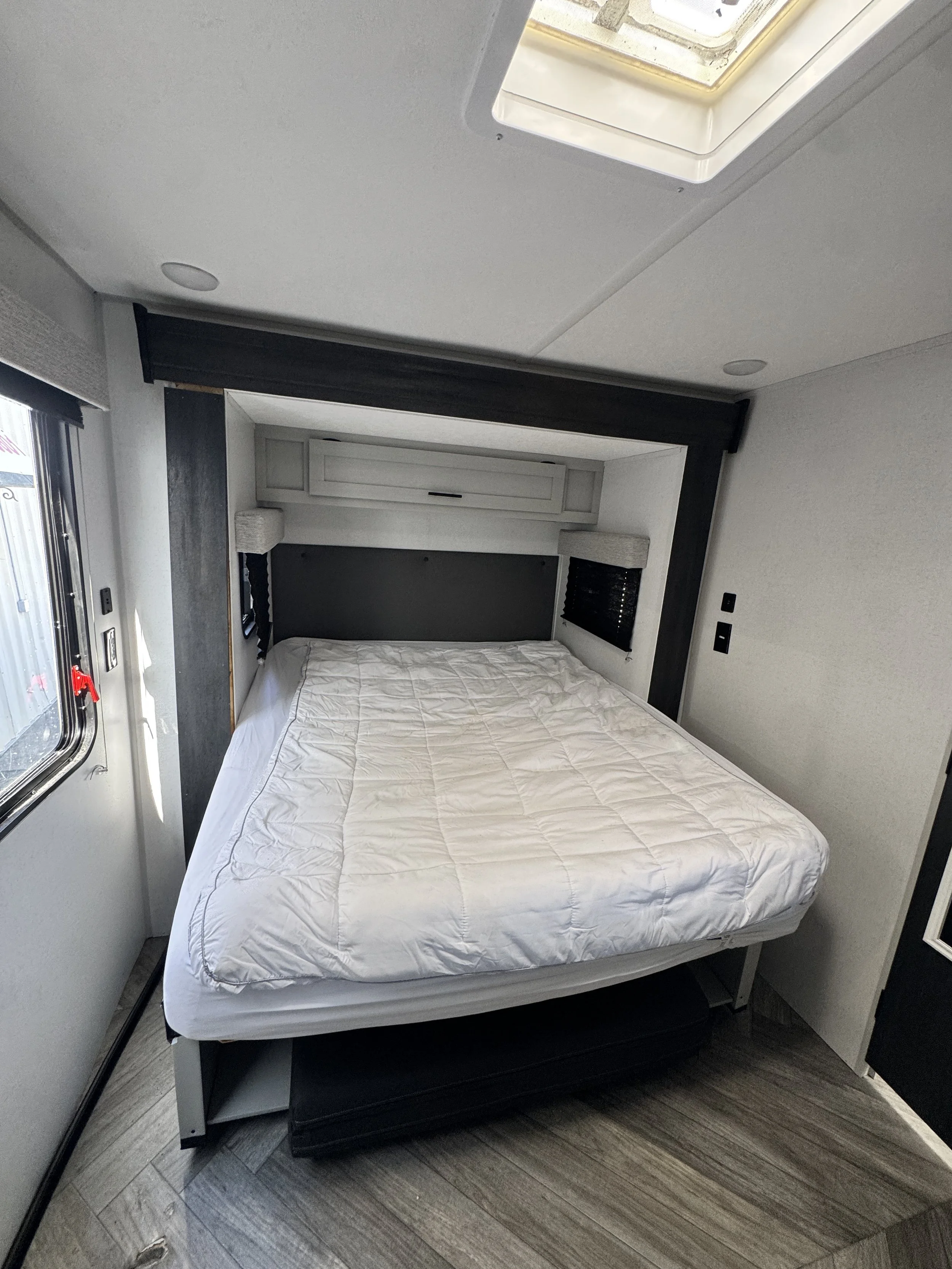 A bedroom with a large bed covered with a white quilt, black window blinds, a window with a partially open blind, and overhead lighting in a modern RV interior.