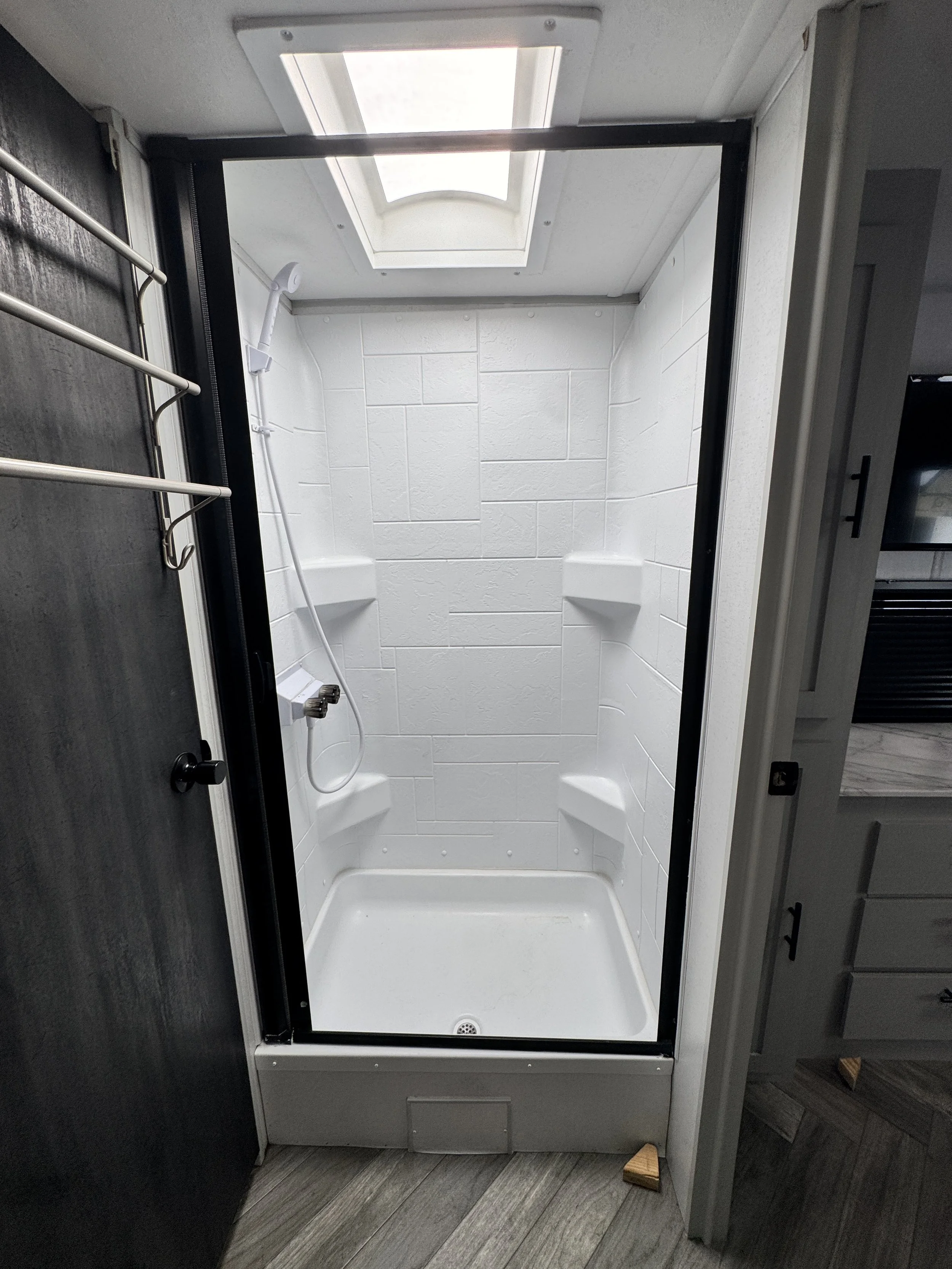 Small shower stall with white tiled walls, a skylight window above, a handheld showerhead, and built-in corner shelves, enclosed in a black frame.