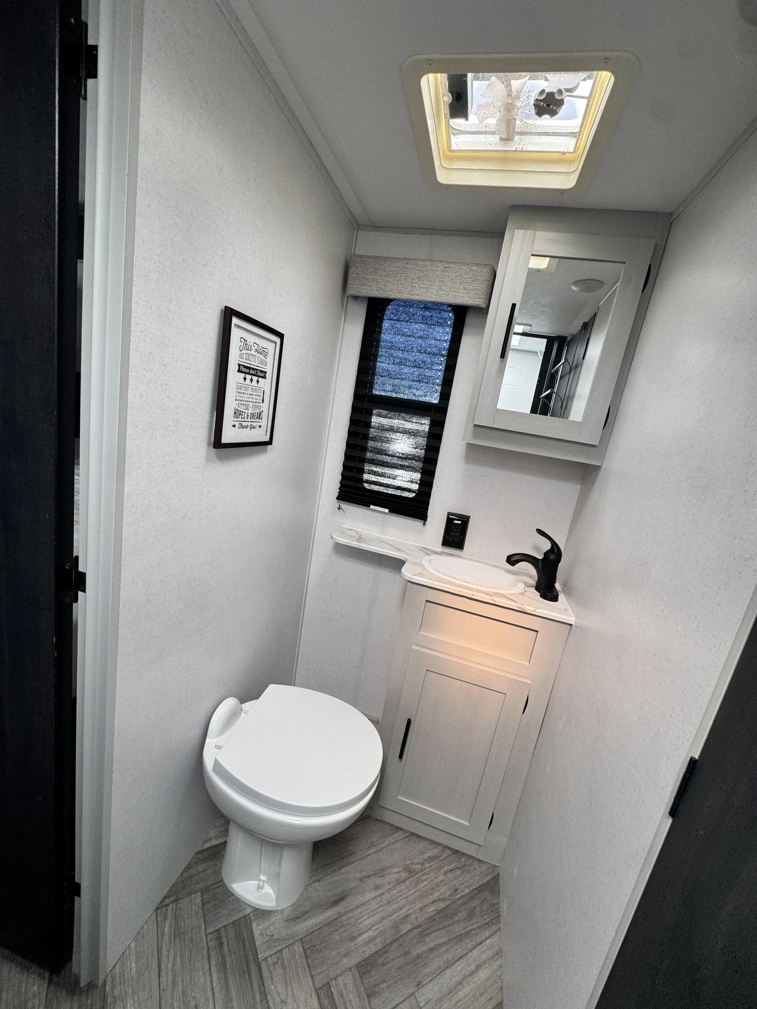 Inside a small bathroom with a white toilet, a cabinet with a mirror, a window with a black blind, and a small decorative picture on the wall.