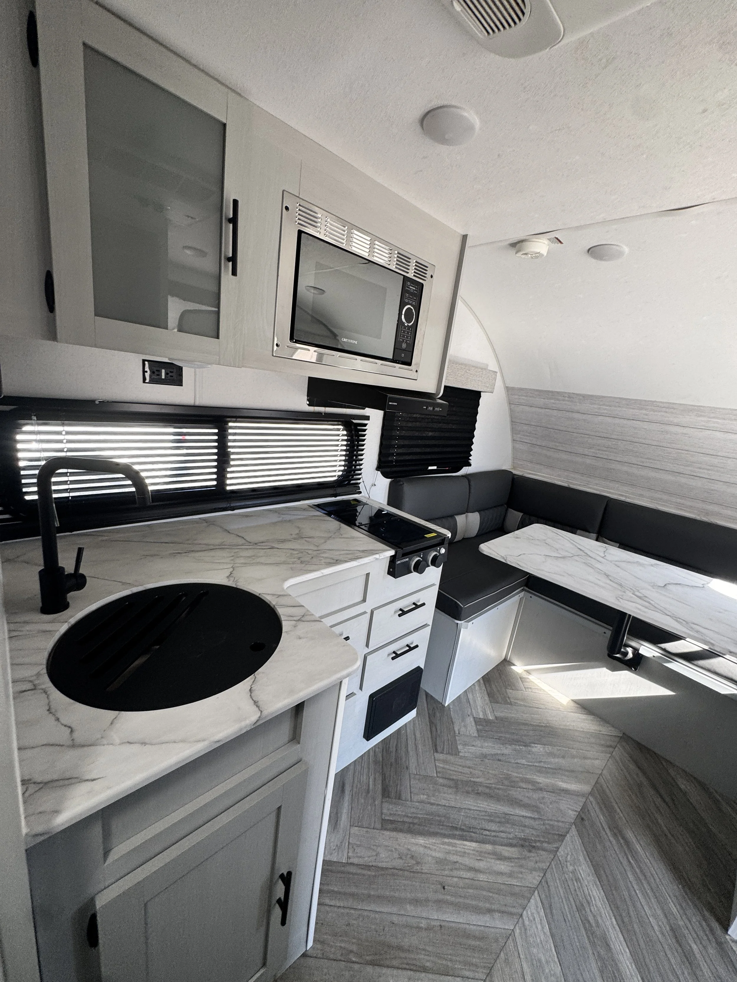 Kitchen area with a white marble countertop, black sink faucet, microwave oven built into a white cabinet, and a black cabinet with window blinds in the background. The floor has a herringbone pattern with gray wood planks.