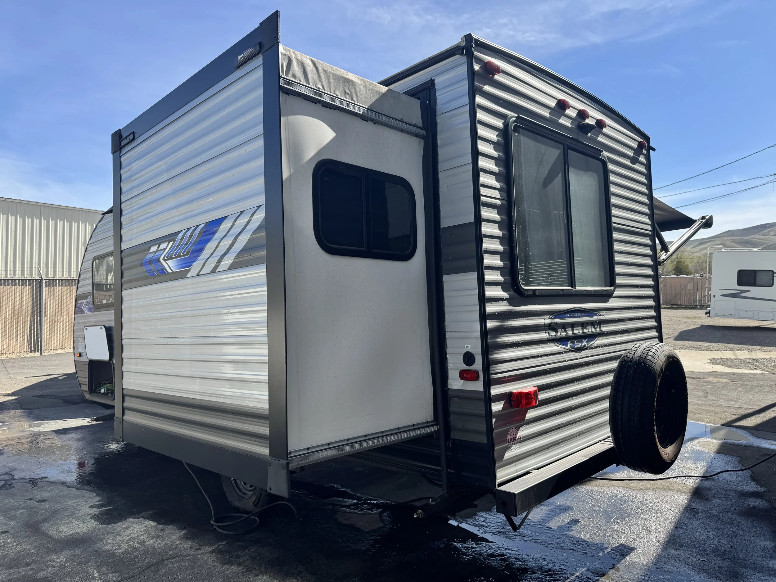 A silver travel trailer with a black door and window, parked on an outdoor lot. The trailer has horizontal stripes and branding on the side. The sky is clear and blue, with mountains in the background.
