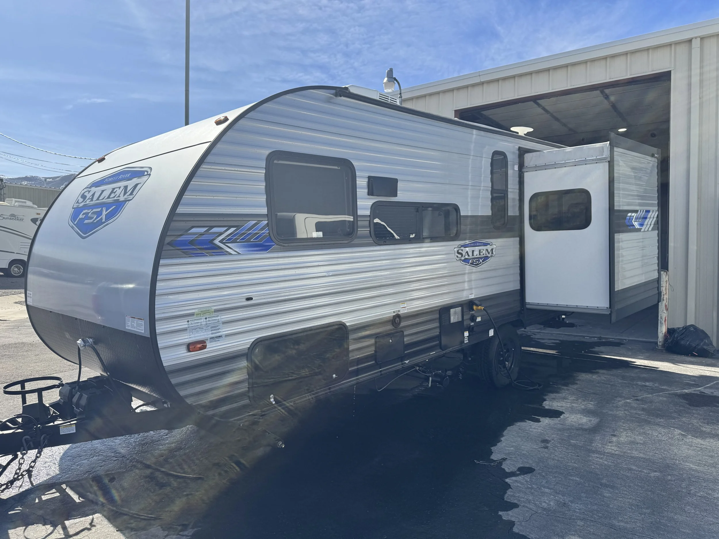 A travel trailer labeled 'Salem FSX' parked outside a building on a sunny day with a clear blue sky. The trailer has windows and an awning, with a hitch at the front.
