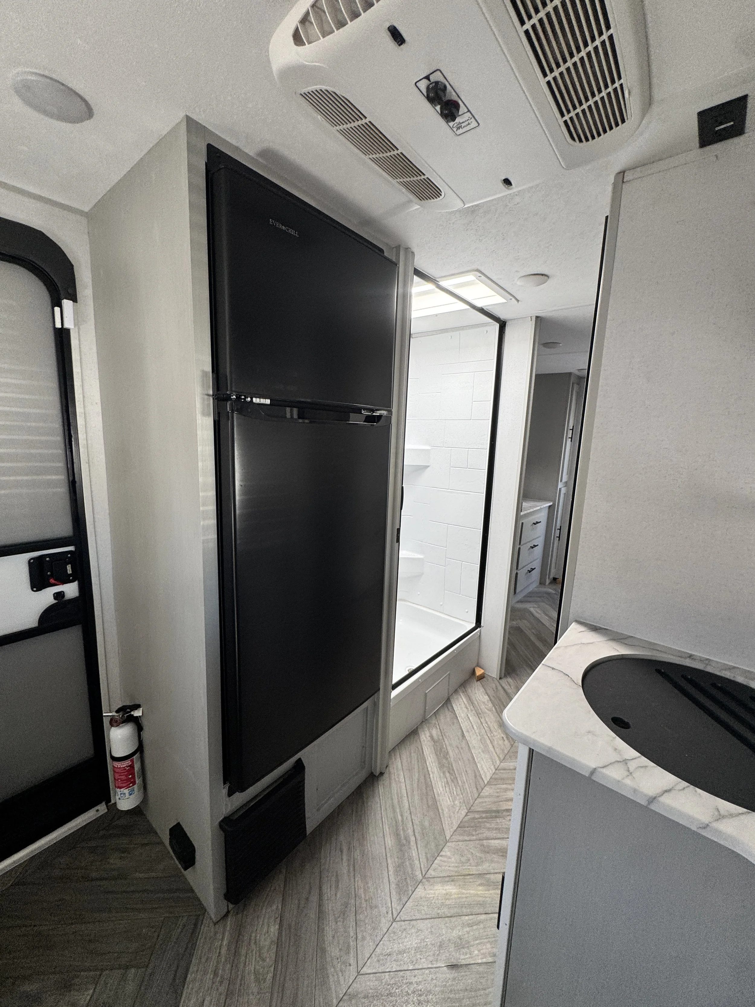 Interior of a small RV or camper with a black refrigerator, a small sink with a black cover, overhead air conditioning unit, and wooden patterned floor.