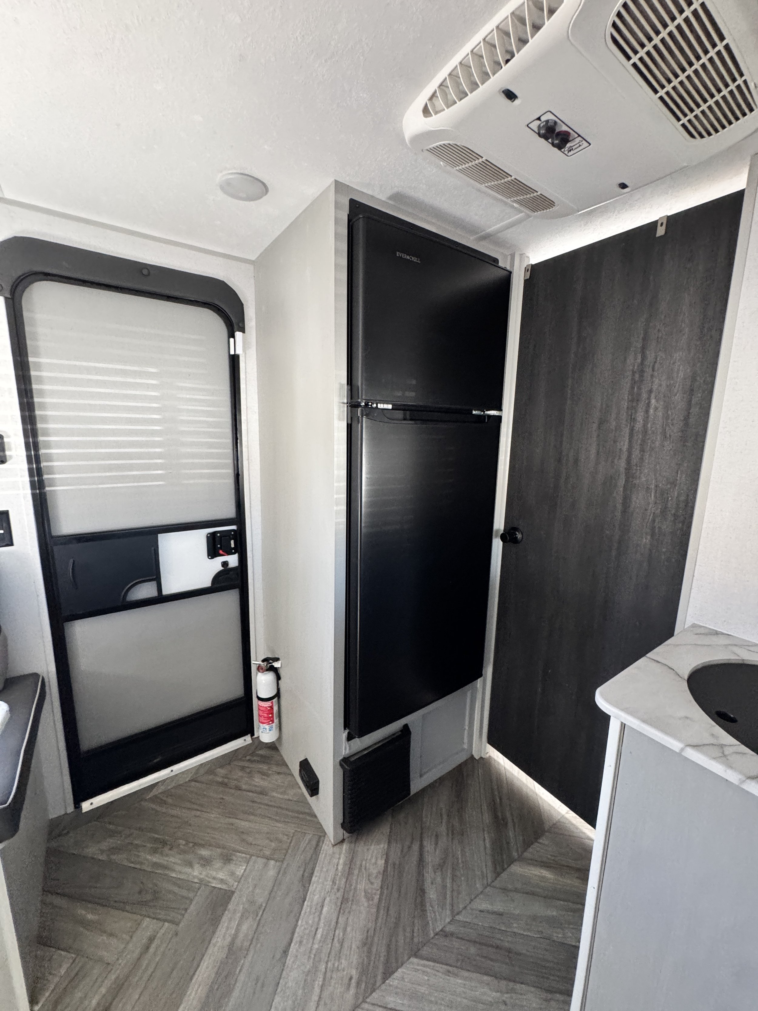 Interior of a small recreational vehicle or camper with a black mini refrigerator, a white and black cabinet with a fire extinguisher underneath, a small gray table with a built-in sink, and wood laminate flooring.