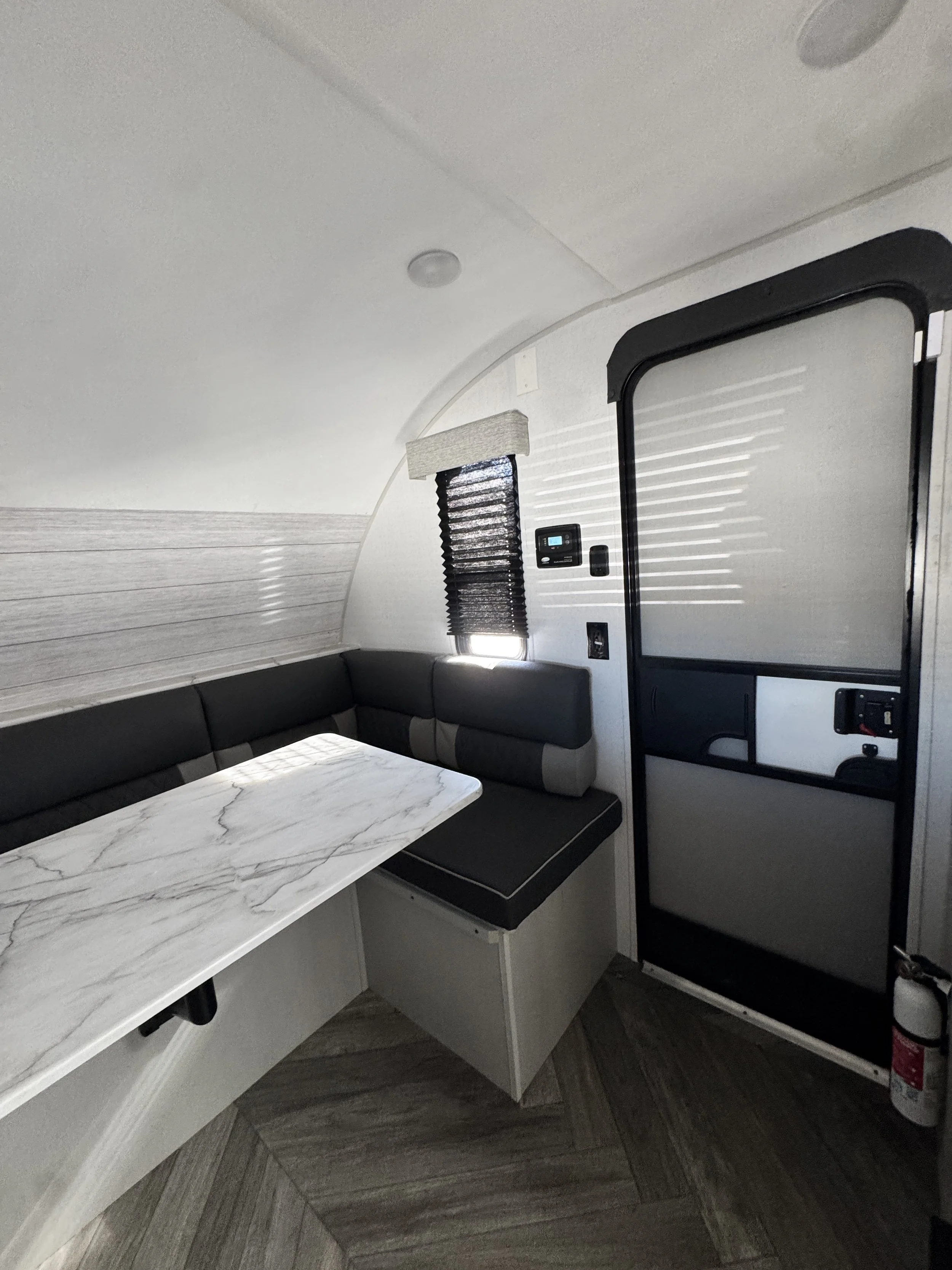 Interior of an RV or camper with a curved ceiling, a marble-like table, black cushioned seating, window with blinds, control panel, and fire extinguisher.