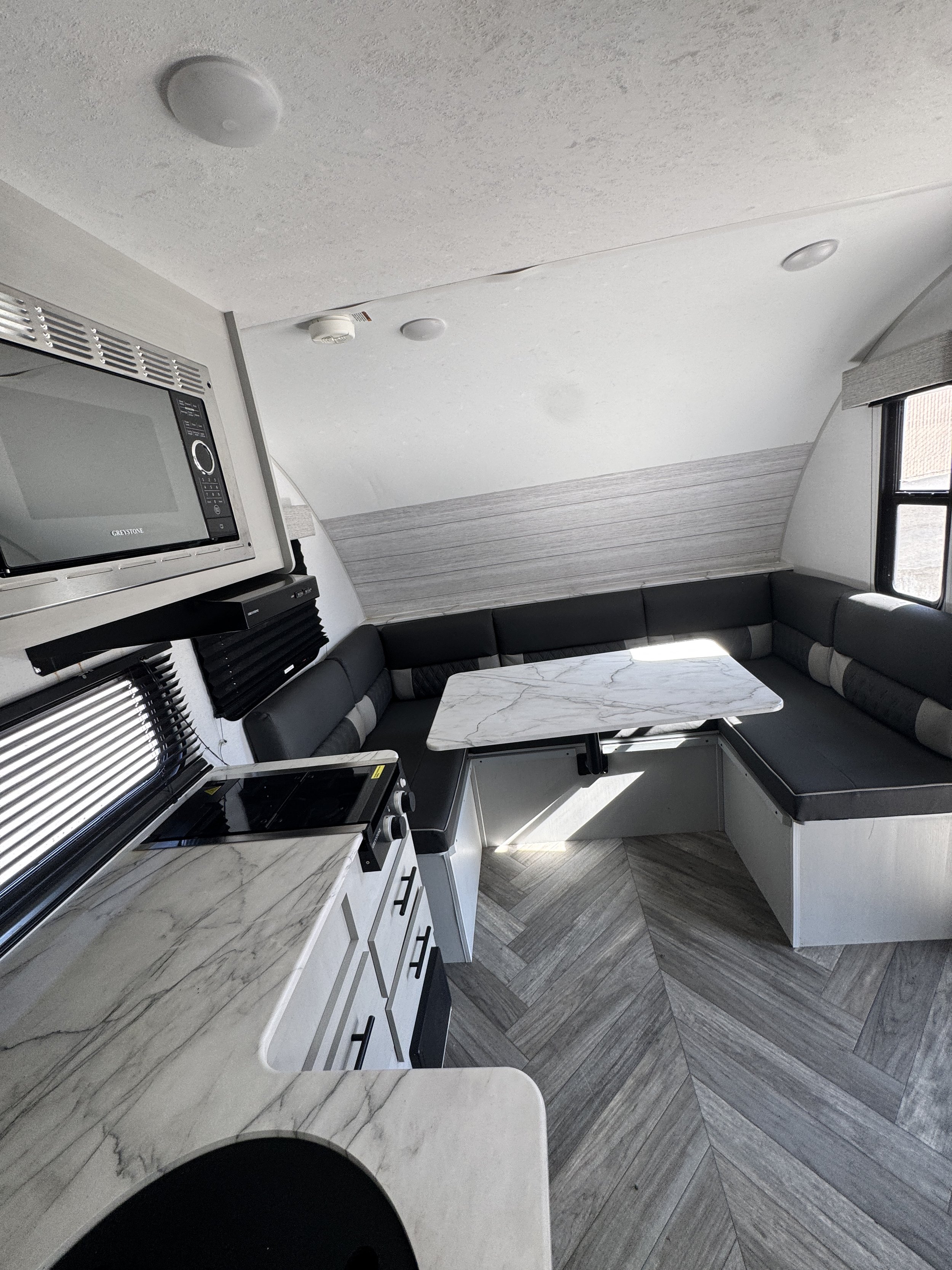 Interior of a compact kitchen and dining area with a curved black sofa and a white marble table, a microwave oven, a window with blinds, and a patterned floor.