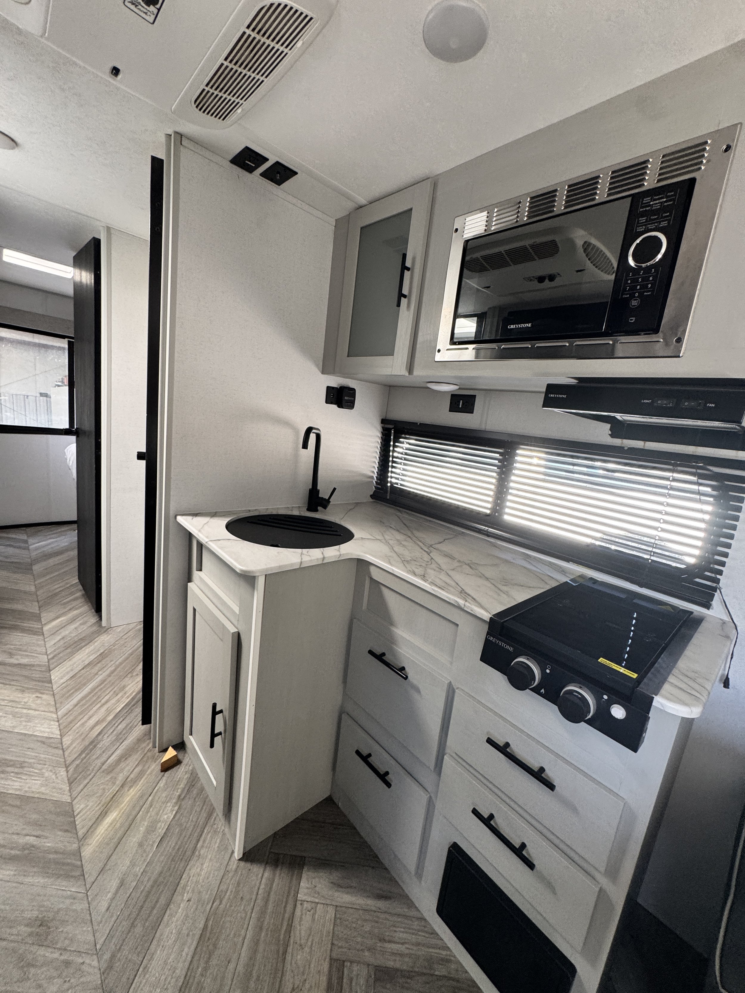 A modern kitchen with a white marble countertop, black faucet, upper gray cabinets with glass doors, a microwave, a stove, and a window with black blinds.