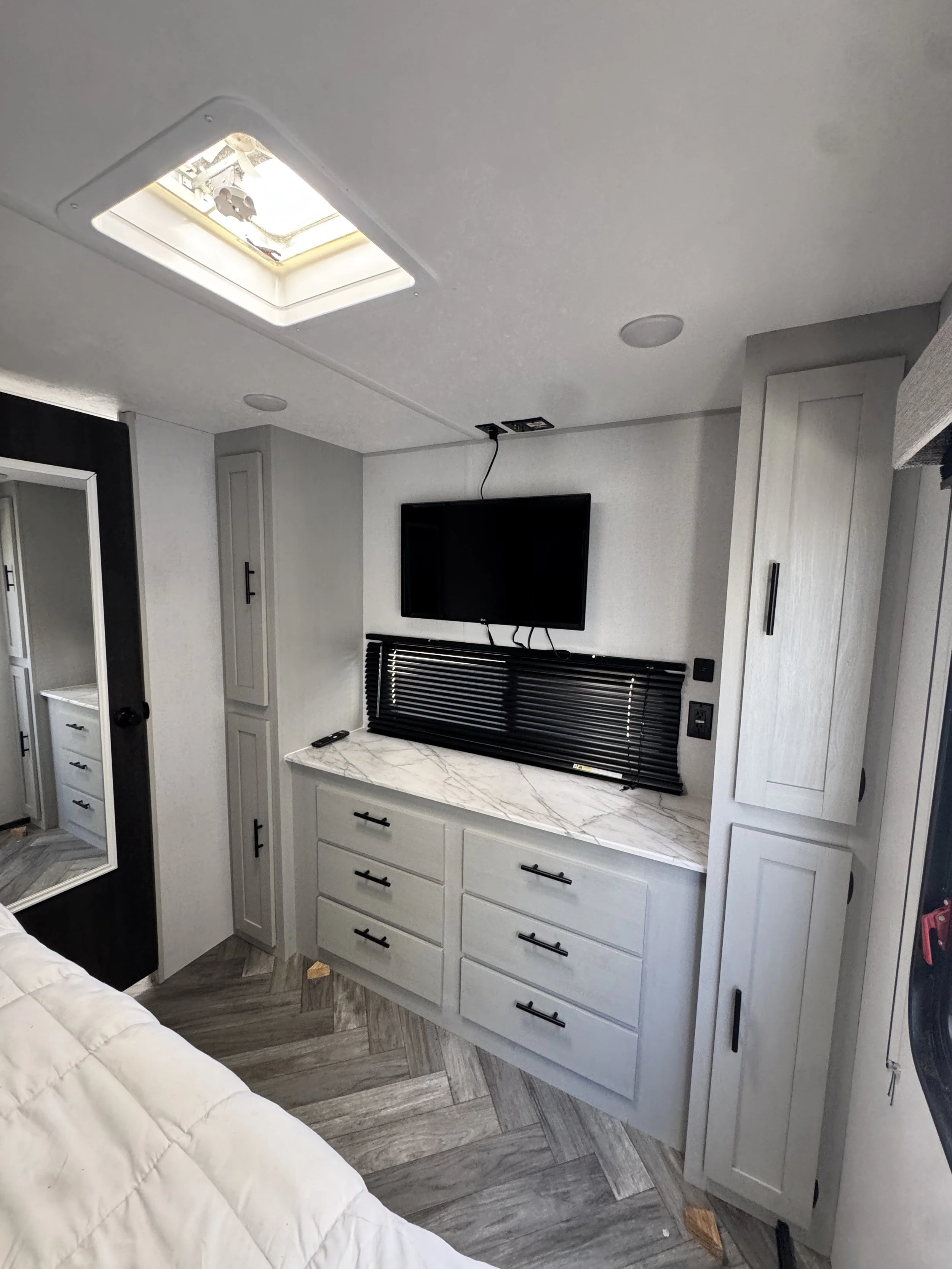 The interior of a modern recreational vehicle or camper with light-colored cabinetry, a mounted flat-screen TV, a marble countertop, and a window with black blinds. There is a ceiling vent and wood-patterned flooring.