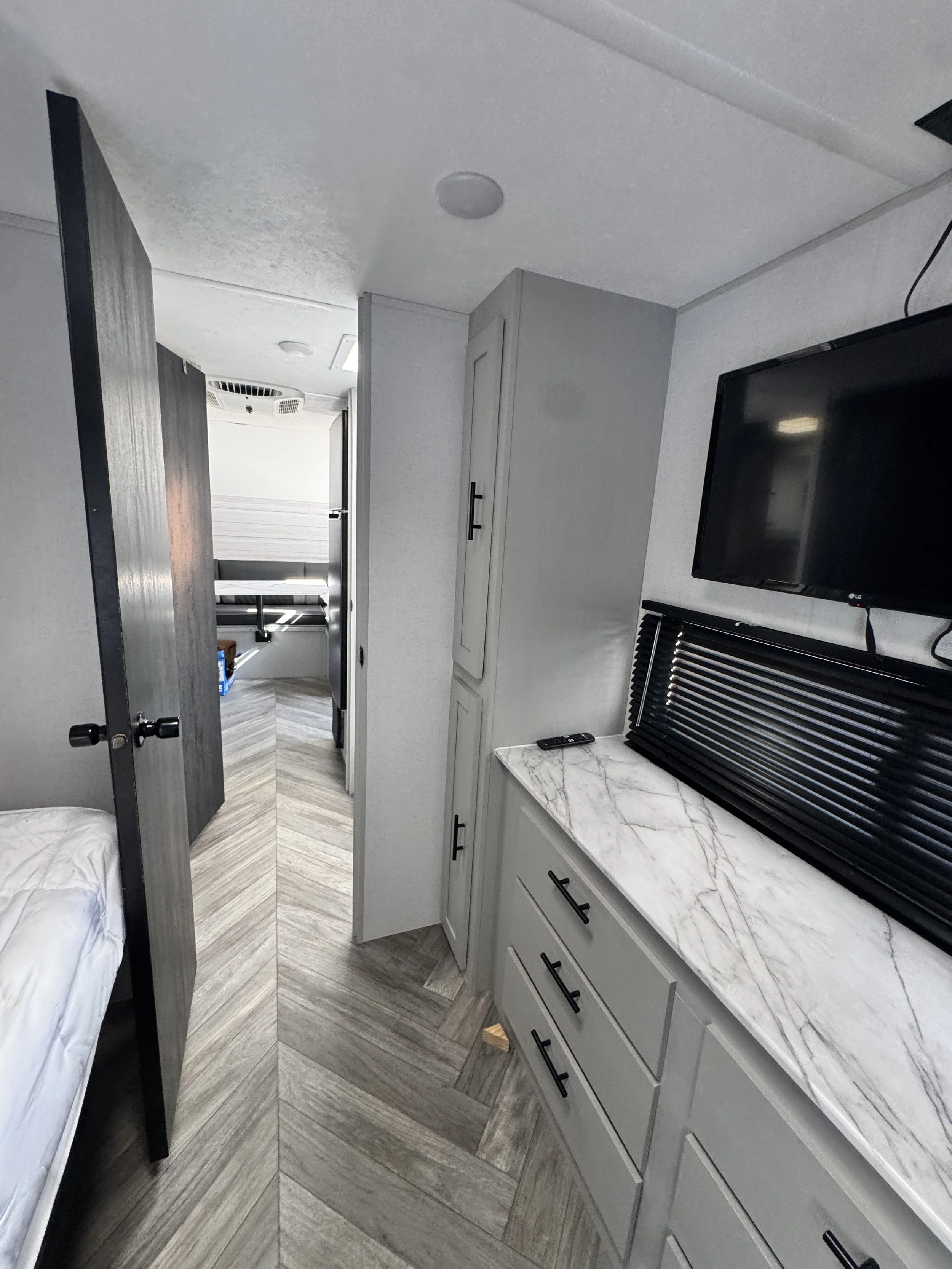 Interior view of a small room with a bed, a dresser with a marble countertop, a wall-mounted TV, and a hallway leading to another room with a bed and window.