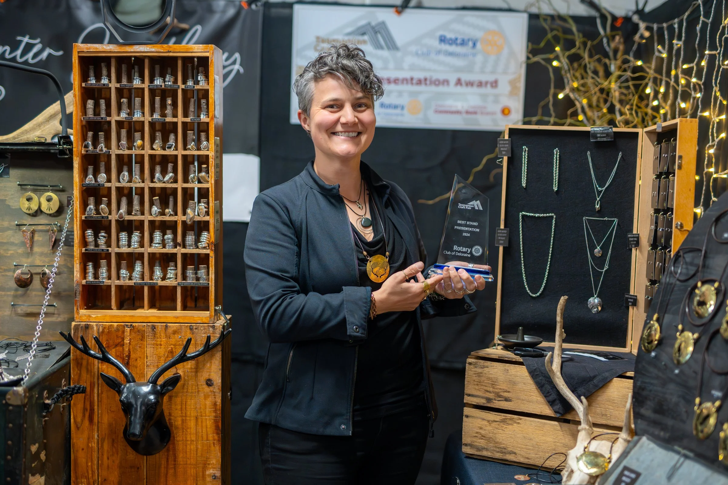 A smiling woman holding an award at a jewelry display booth with necklaces, earrings, and jewelry pieces on display, and a deer head sculpture on a wooden stand.