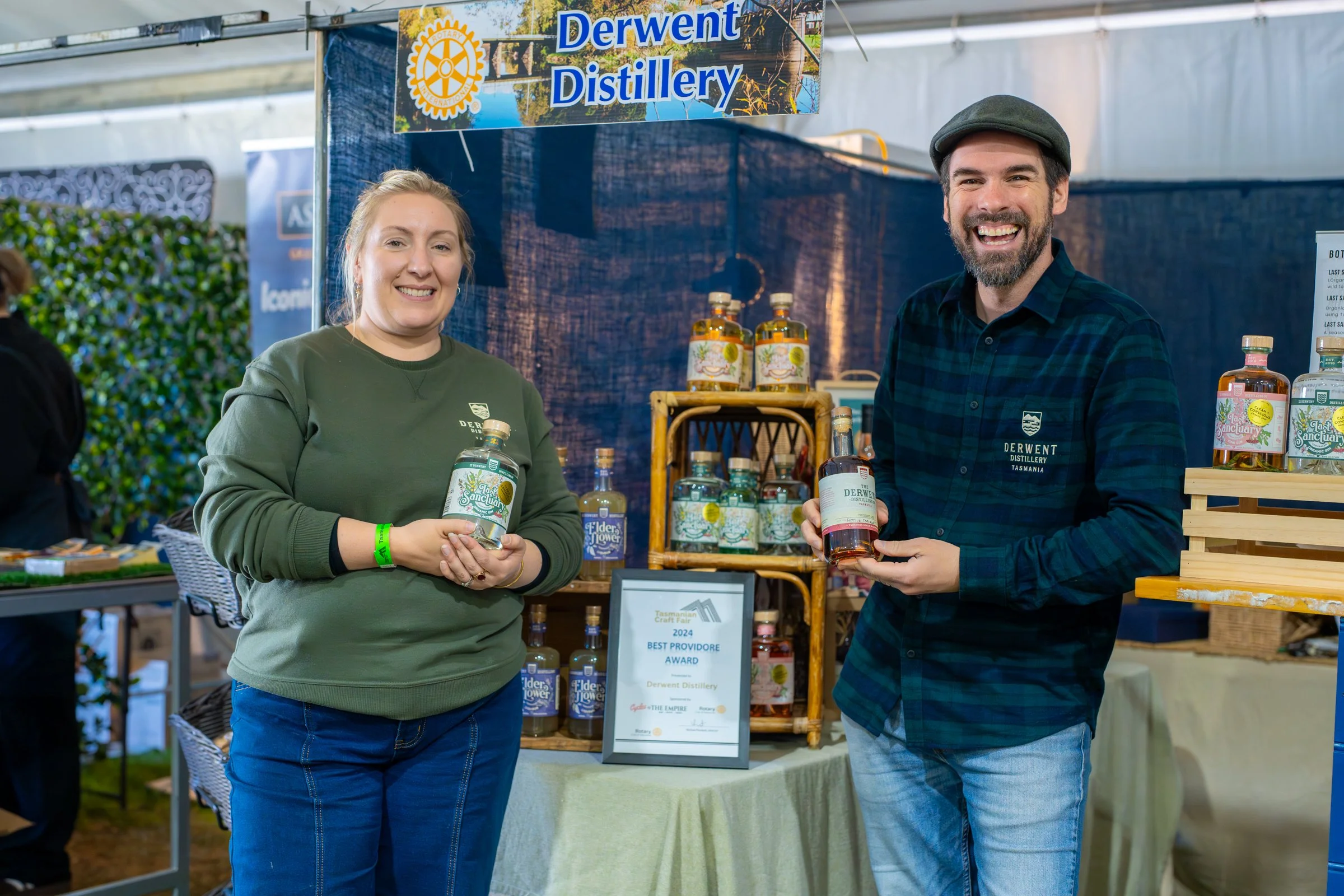 Two people smiling at a booth labeled 'Derwent Distillery' at a fair or market. They are holding bottles of distilled products, with shelves of bottles and a framed award on display behind them.