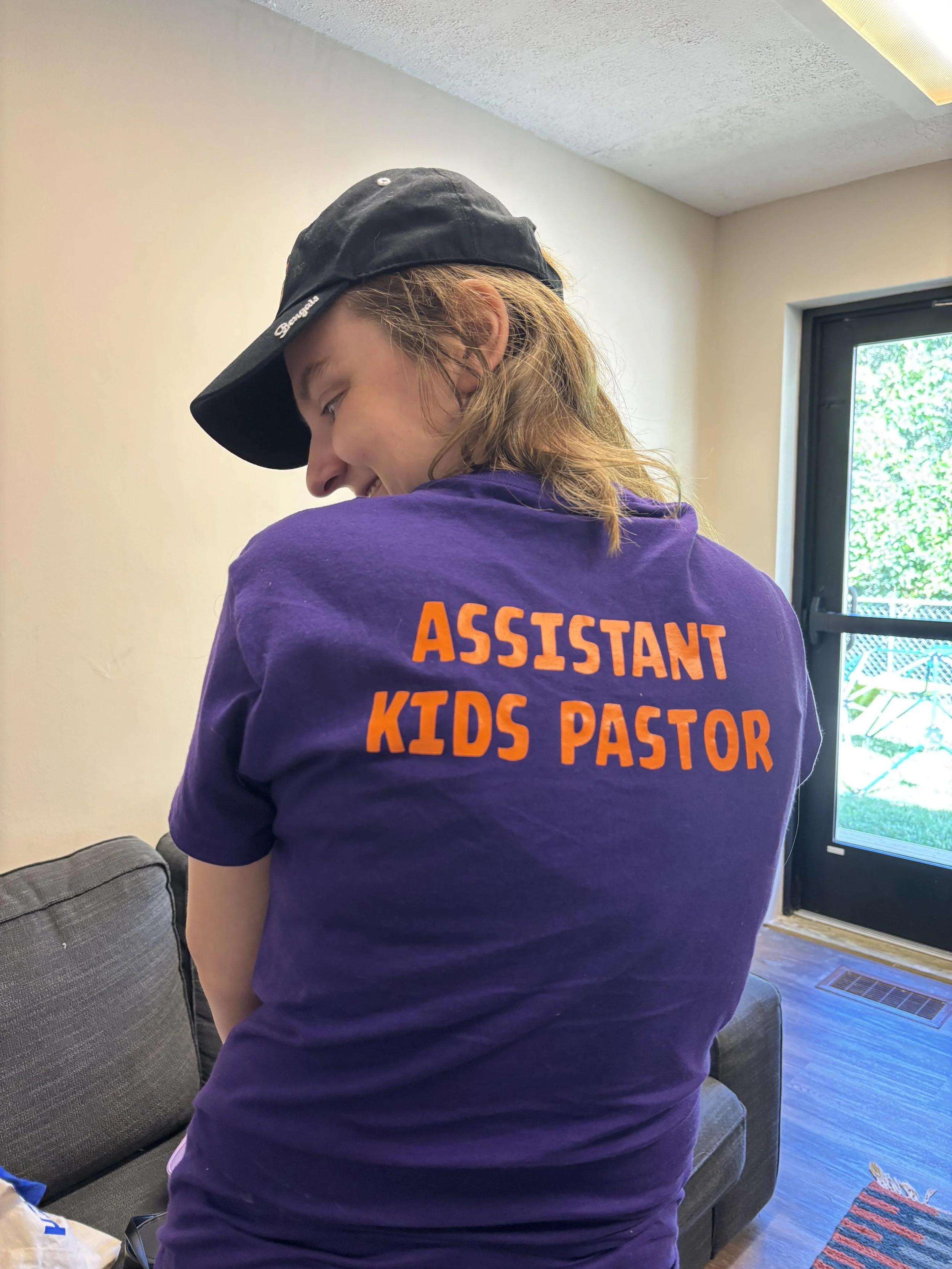 A young woman with brown hair wearing a black baseball cap and a purple T-shirt that says 'Assistant Kids Pastor' in orange text, standing inside a room near a window.