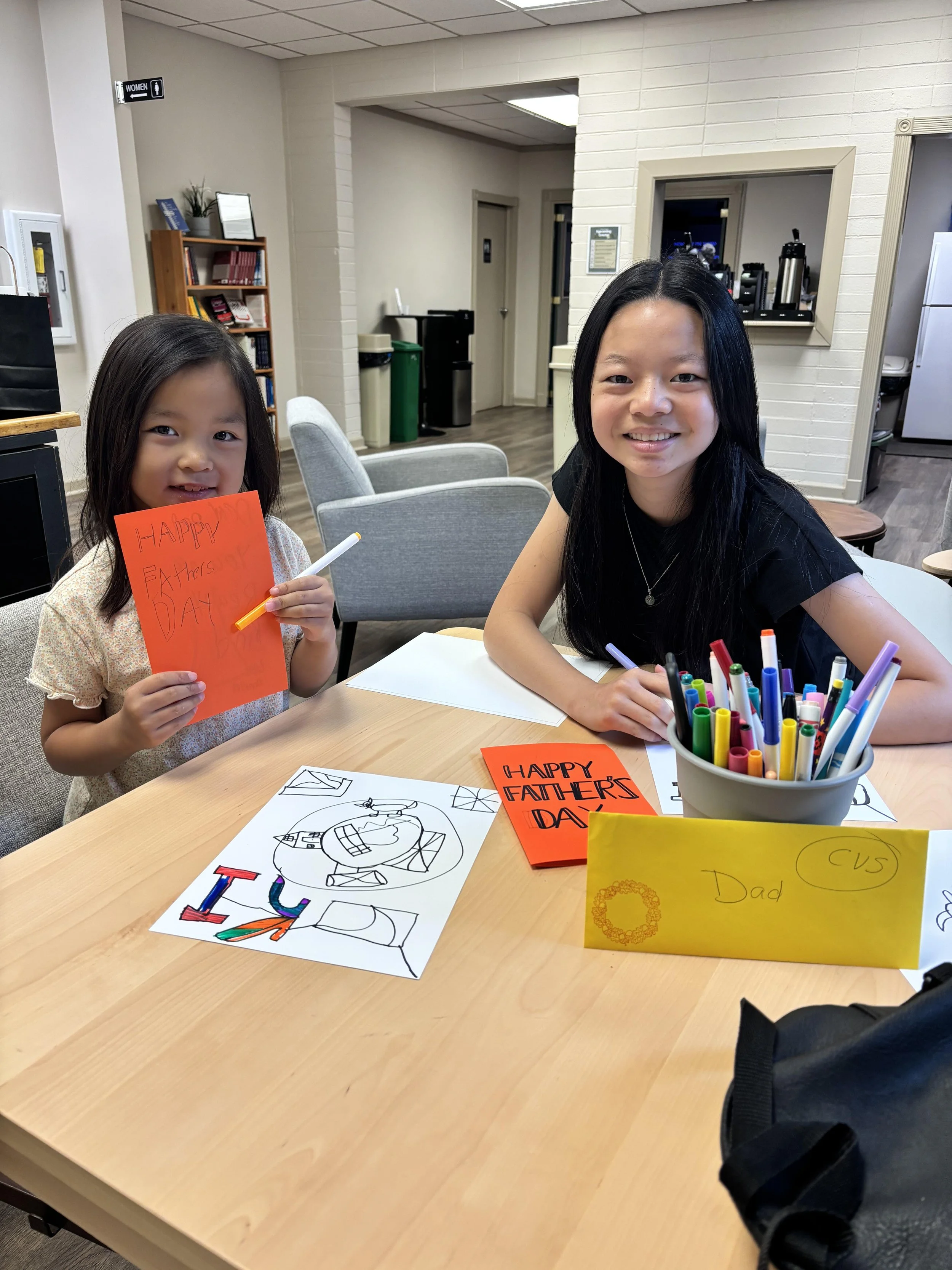 Two girls sitting at a table with colorful markers and craft projects, holding greeting cards for Father's Day. The girl on the left holds an orange card that says 'HAPPY Father's DAY,' and the girl on the right has a yellow card labeled 'Dad.' They are smiling, and there are drawings and crafts on the table.