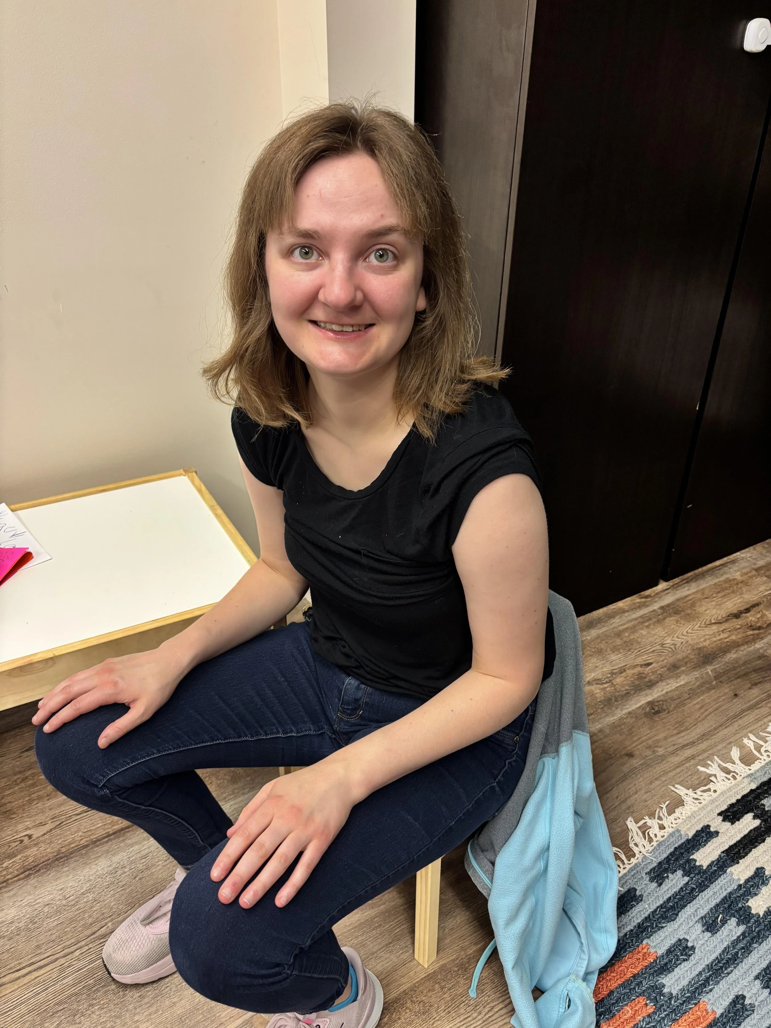 A young woman with shoulder-length light brown hair, wearing a black t-shirt and dark blue jeans, sitting on a chair in a room with wooden flooring, a black cabinet, and a small wooden table.