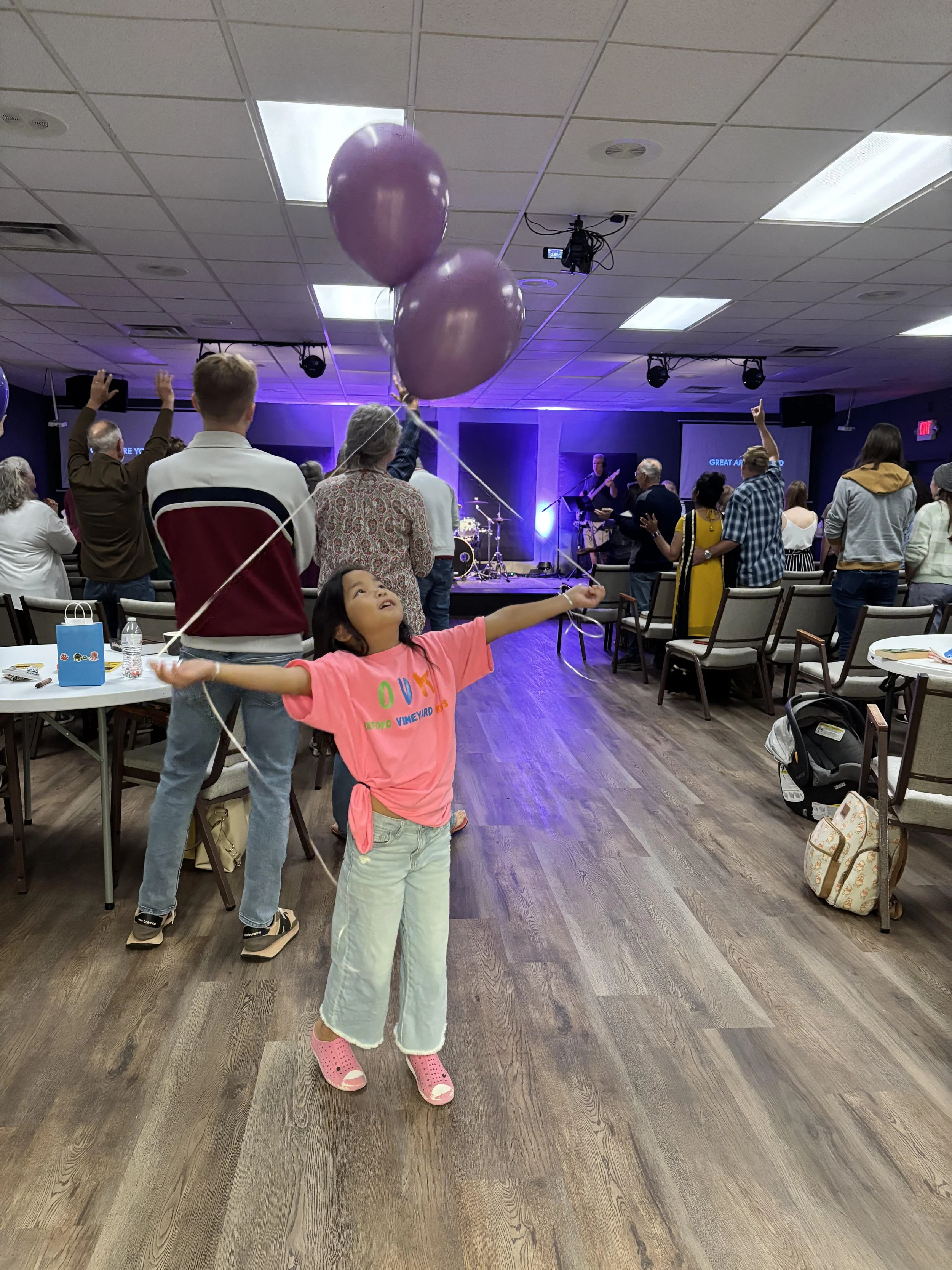 A young girl in a pink shirt and light pants is holding purple balloons in a large room with people attending an event or concert. The stage has musicians playing, with purple lighting, and the room has wooden flooring and tables with chairs.