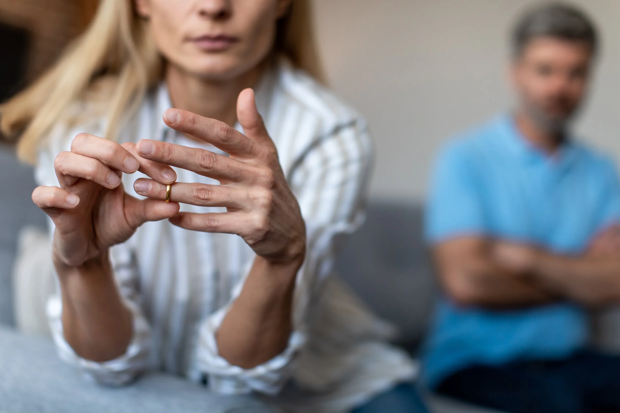 A woman with blonde hair, wearing a white and beige striped shirt, is holding her hands together with a wedding ring visible on her finger. A man with gray hair, wearing a blue shirt, is sitting in the background with his arms crossed, out of focus.