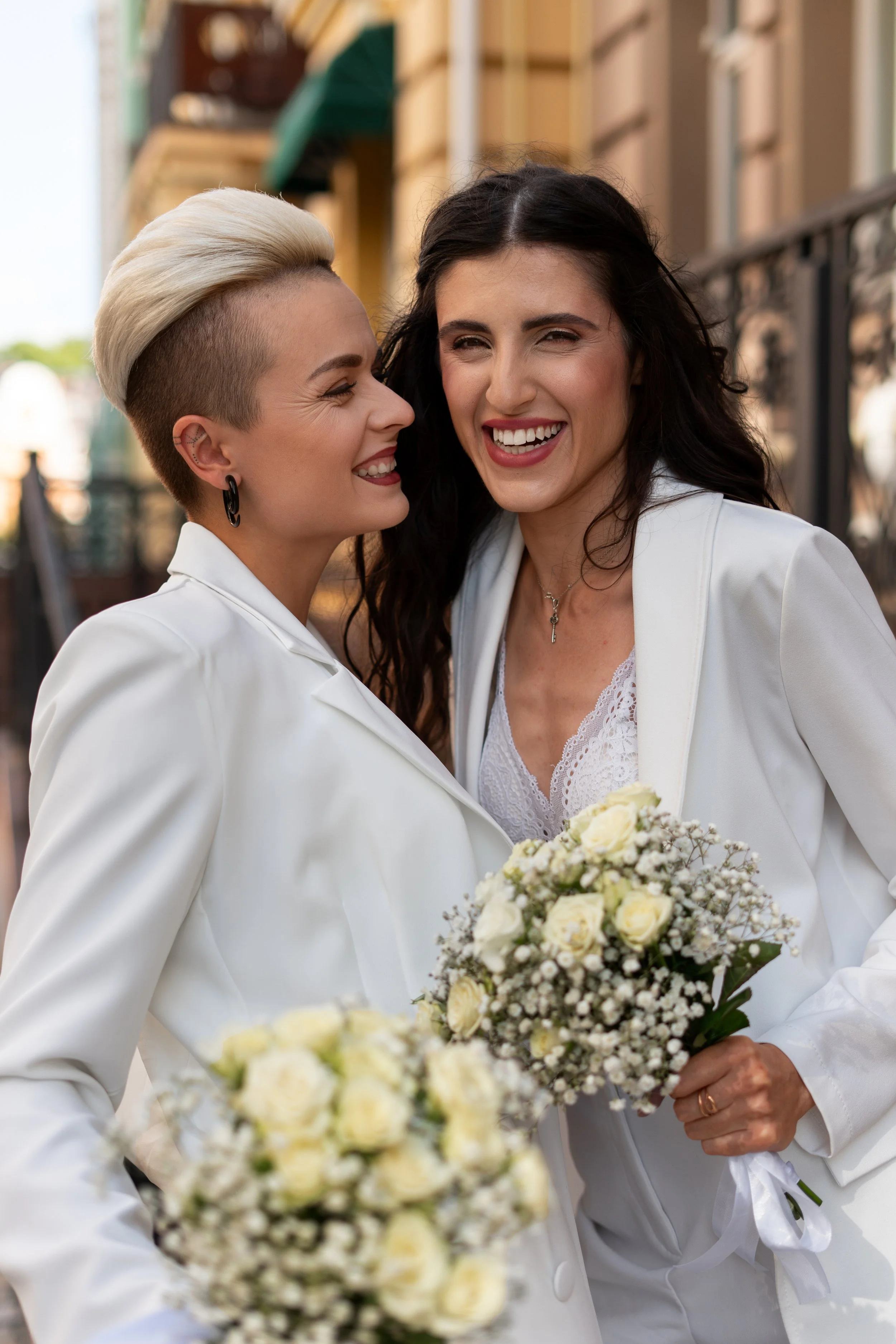 Two women in white suits smiling and embracing each other outdoors, holding bouquets of white roses and babys breath.