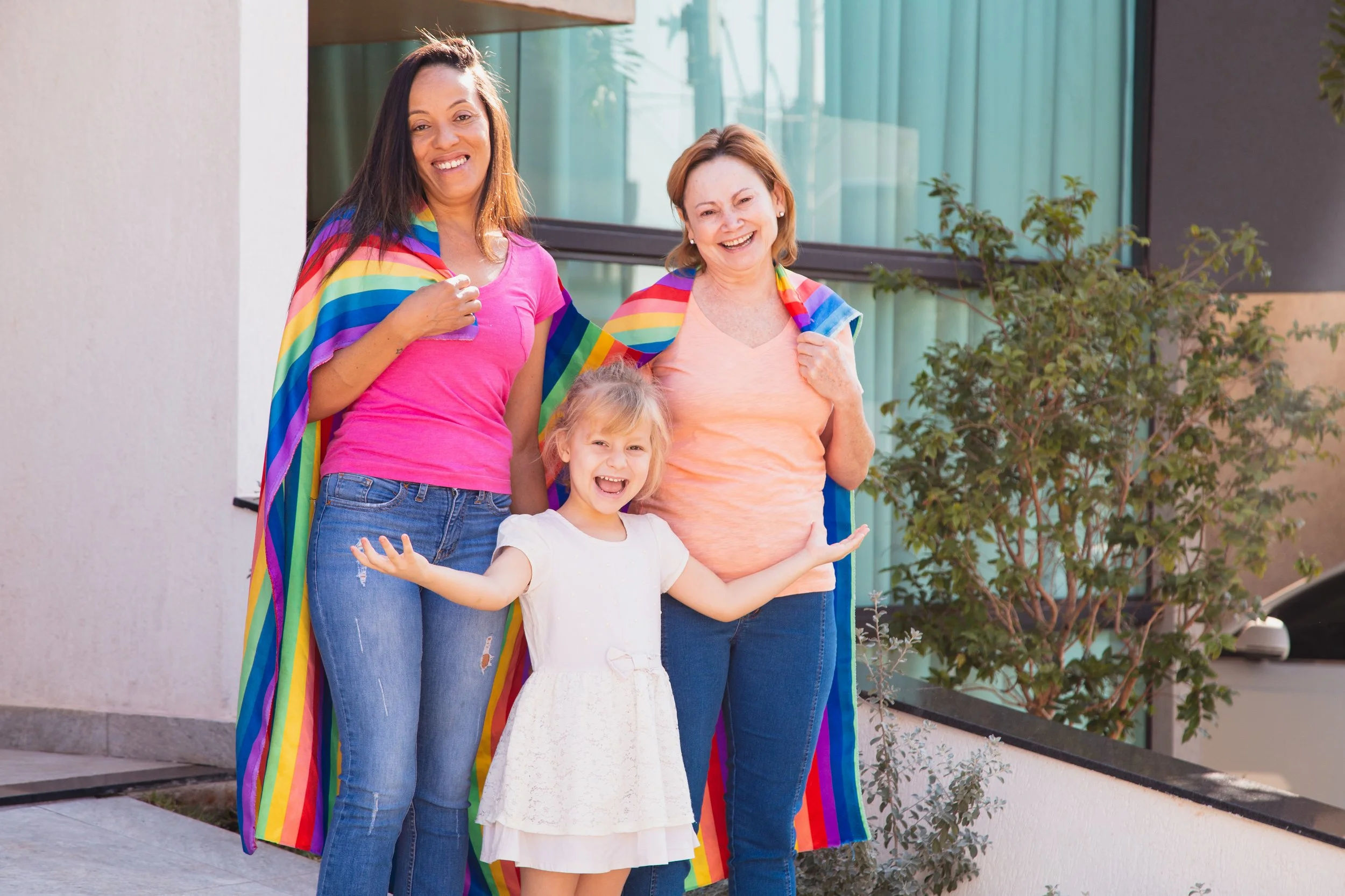 Three women and a young girl standing outside in front of a modern building with large windows, smiling and wearing rainbow pride flags draped over their shoulders on a sunny day.