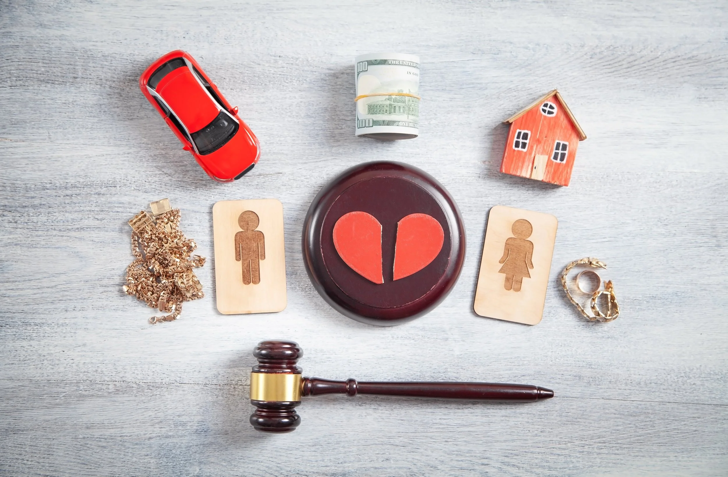 A diagram depicting a broken heart in the center, surrounded by various objects including a red toy car, a rolled bundle of 100-dollar bills, a small wooden house, a pile of gold jewelry, two wooden gender symbols, a gold ring with a stone, and a gavel.