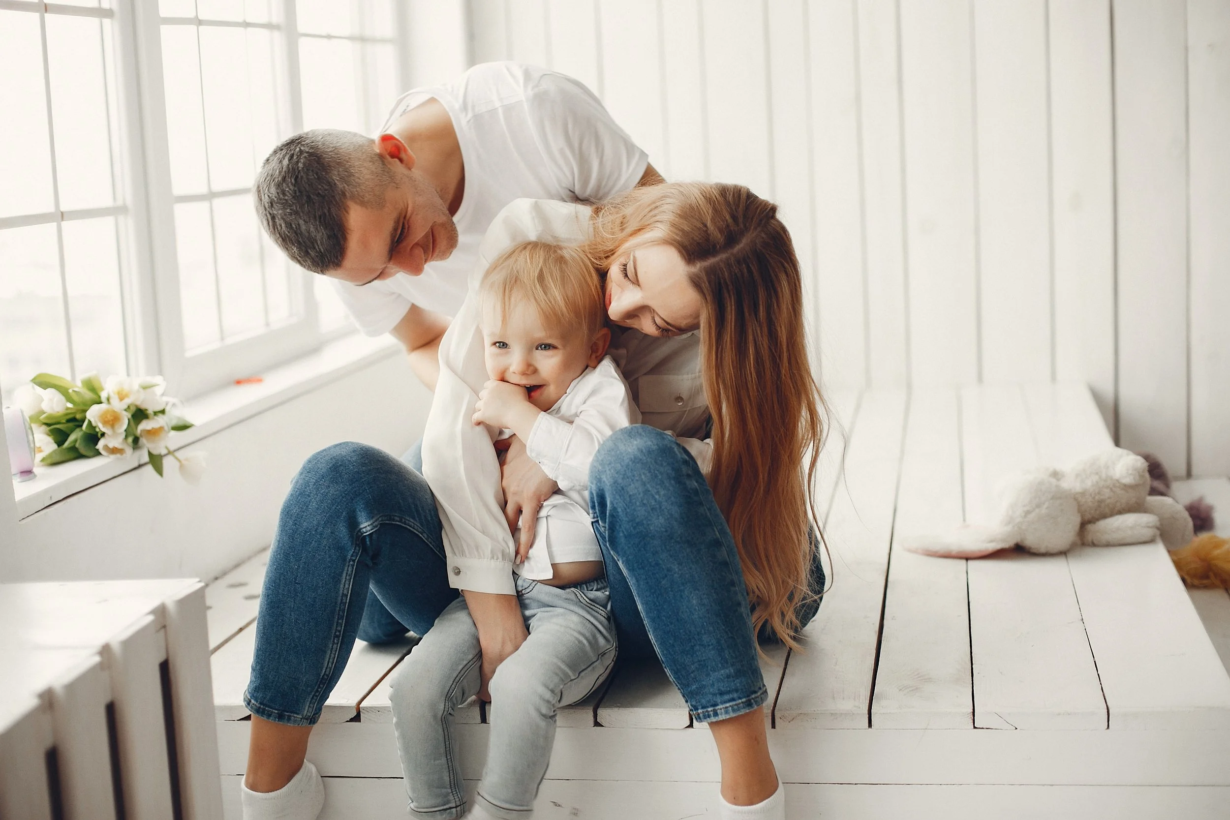 A family of three, a man, woman, and young boy, sitting on a white bench near a window, with white walls and plants nearby. The woman and boy are smiling and cuddling, while the man leans in to join the moment.