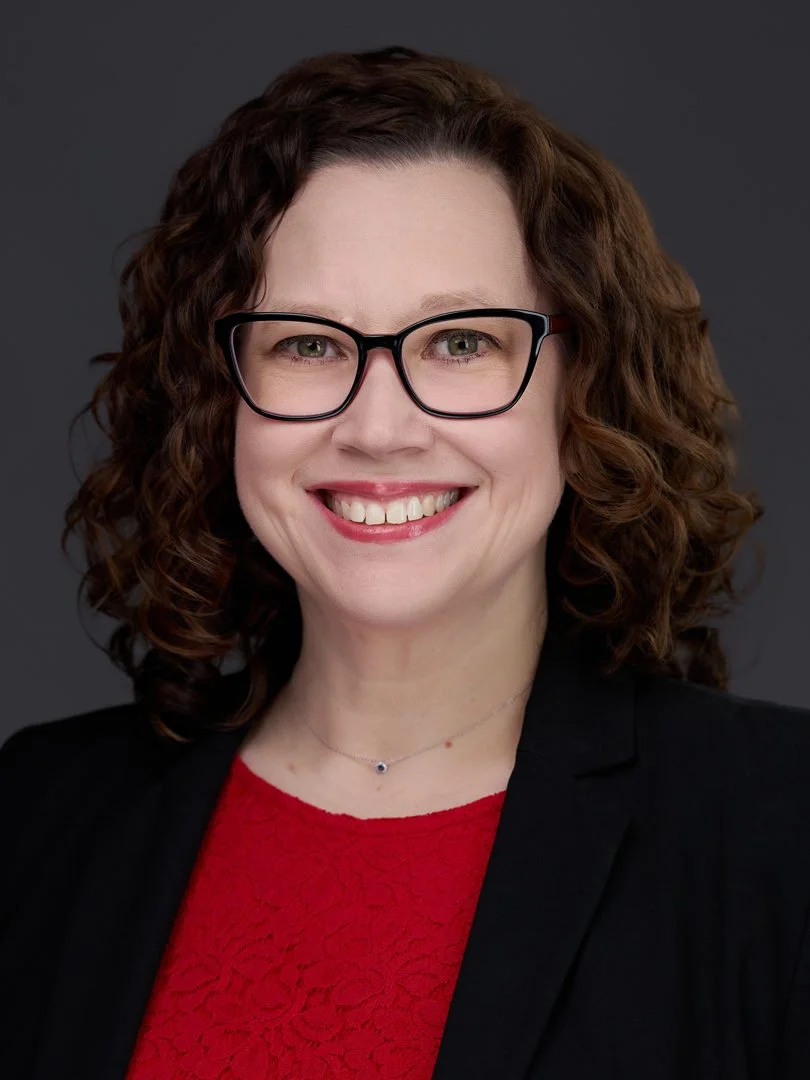 A woman with curly brown hair, glasses, and a bright smile, wearing a black blazer over a red lace top, against a dark background.