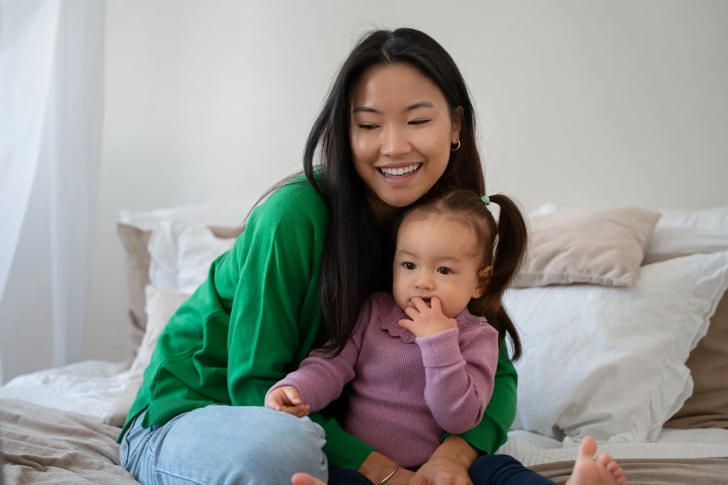 A woman with long dark hair and a green sweater smiling while sitting on a bed with a young girl with pigtails and a pink sweater.