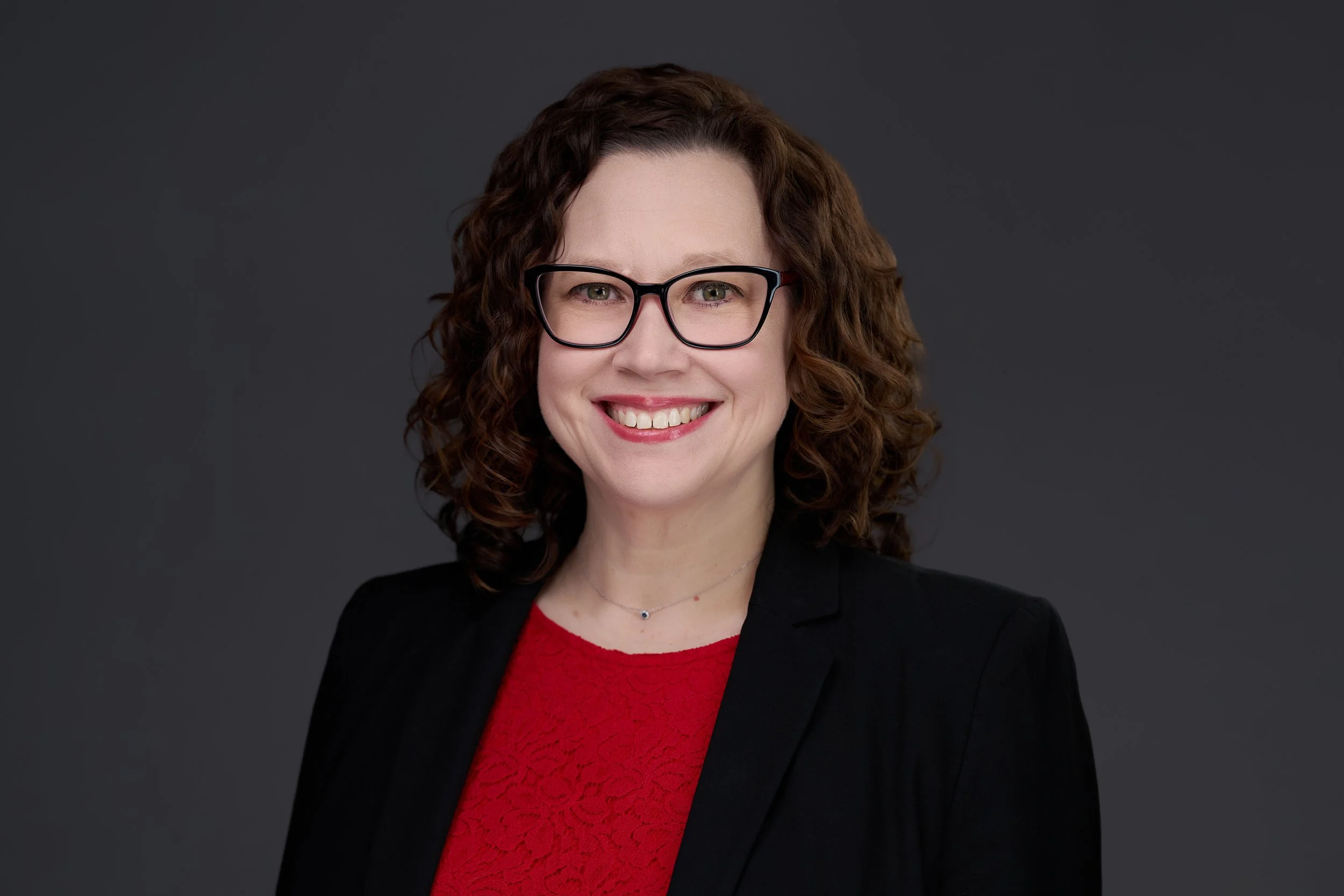 A woman with curly brown hair wearing glasses, a black blazer, and a red top, smiling against a dark gray background.