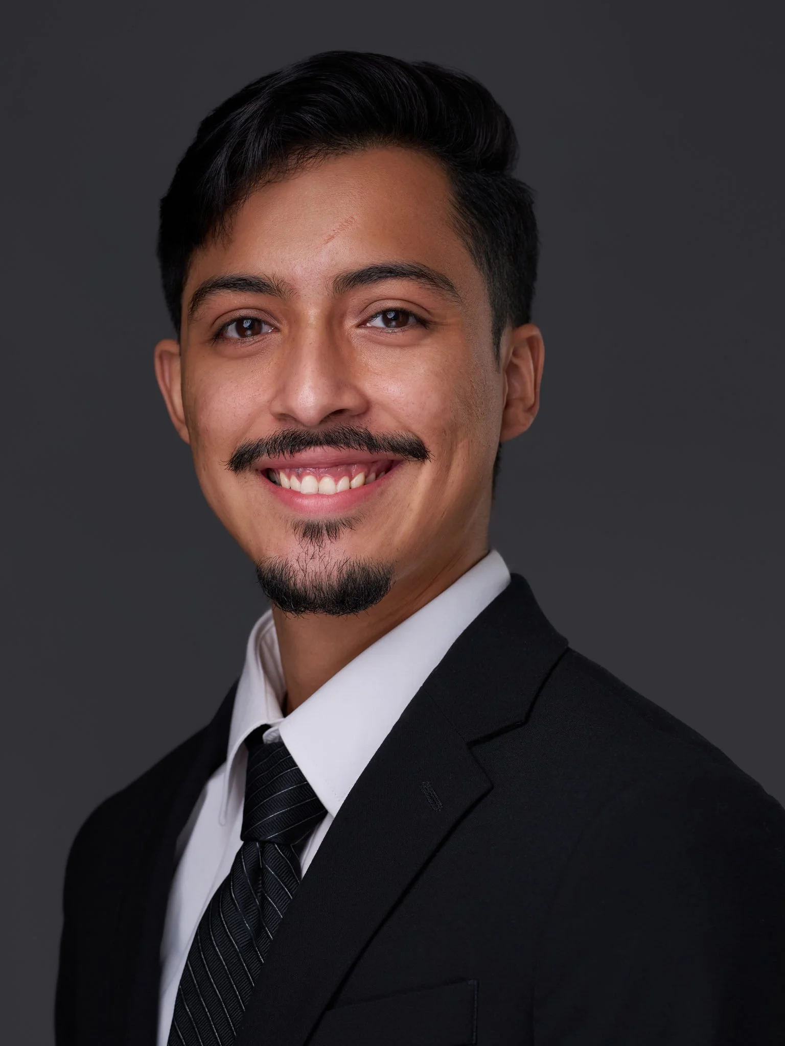 Headshot of a young man with short dark hair, a mustache and goatee, wearing a black suit, white shirt, and black tie against a dark grey background.