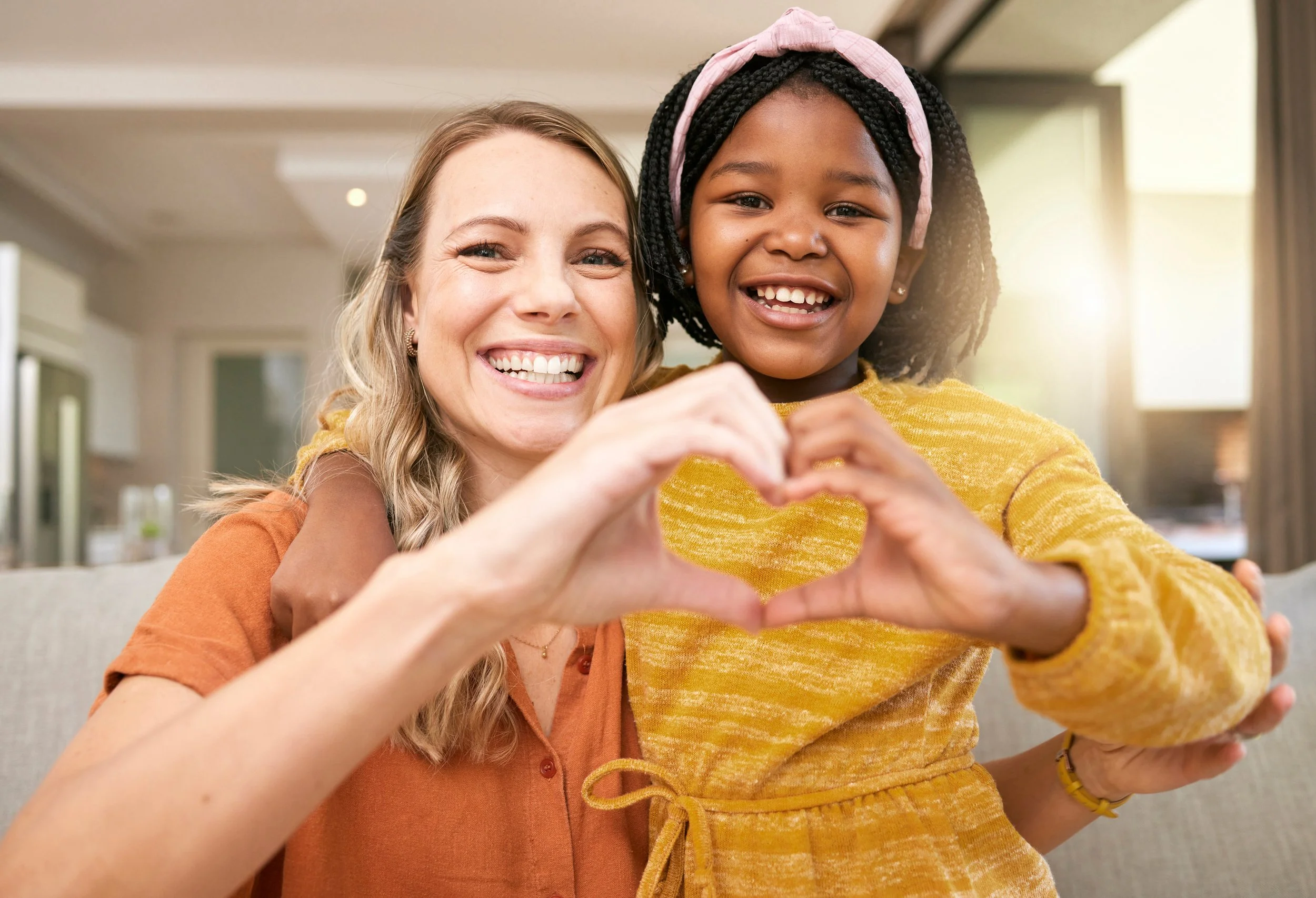 A happy woman and young girl making a heart shape with their hands, smiling inside a well-lit home.