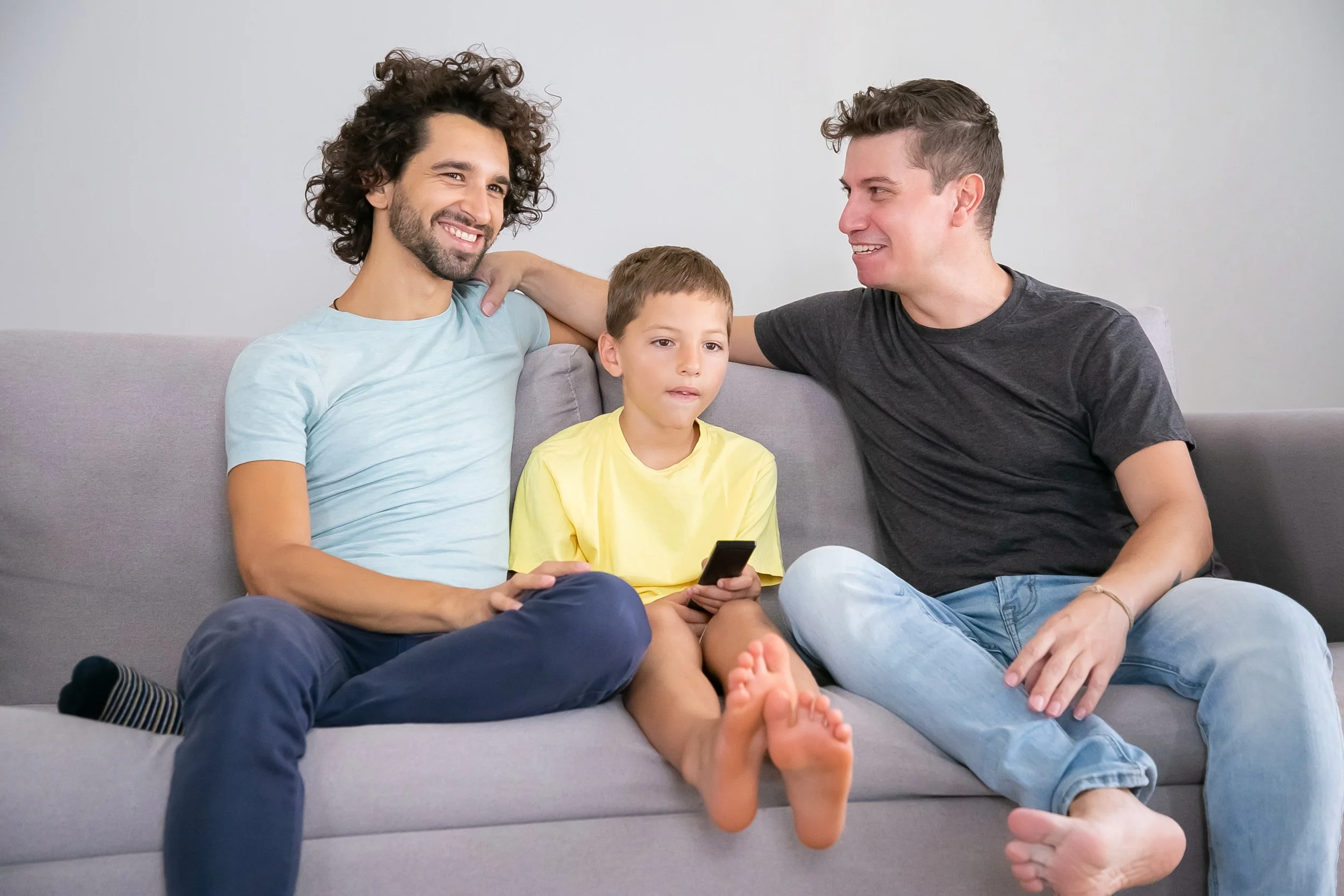 Two adult men and a young boy sitting on a gray sofa, with the boys holding a remote control. The men are smiling and looking at each other, while the boy looks at the camera. The men are wearing casual t-shirts, and the boy is wearing a yellow t-shirt.