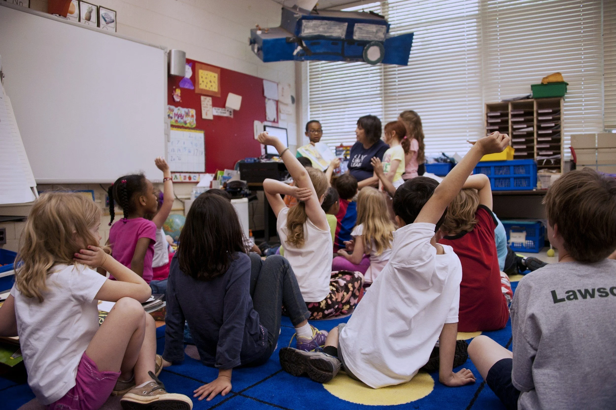 Elementary school classroom with students sitting on a colorful rug, raising hands to answer a question, while a teacher stands near the whiteboard.