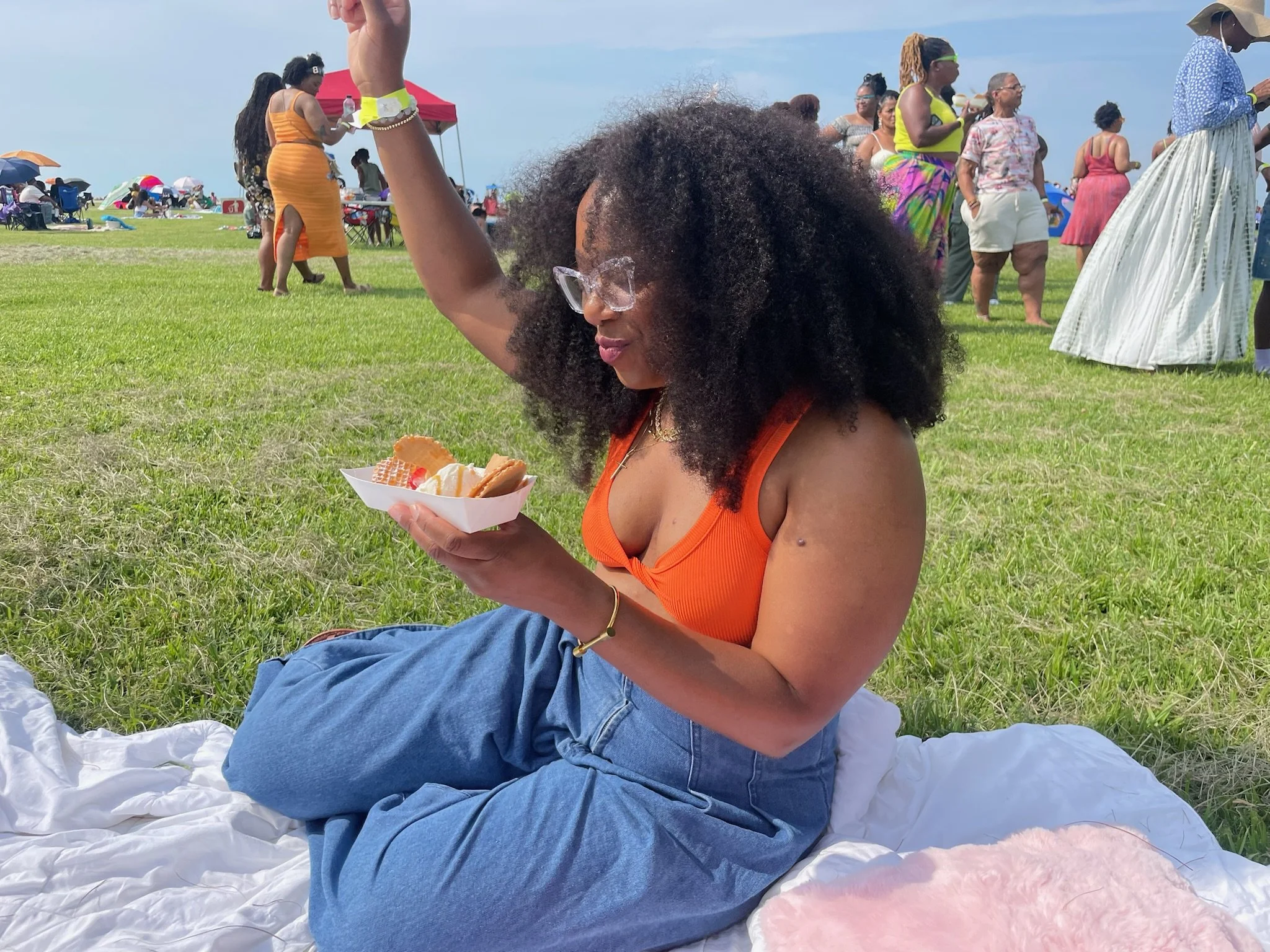 A woman with curly black hair is sitting on a white blanket on the grass at an outdoor gathering. She is holding crazy waffle bar waffles with ice cream, at a lively outdoor event.