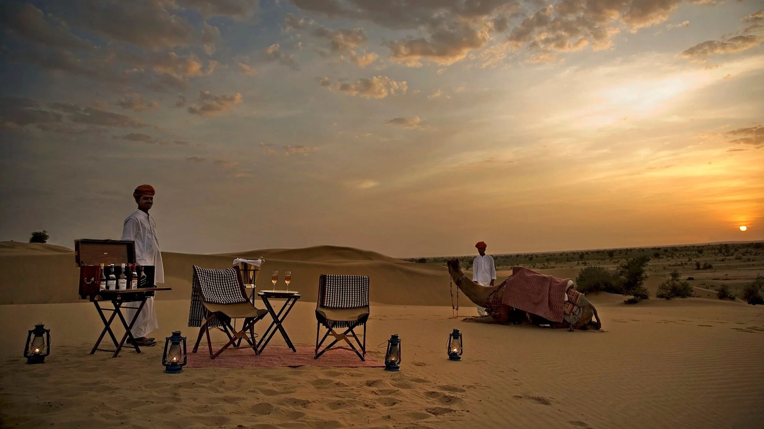Scene of a desert at sunset with two men, one standing with a camel and the other near a table with wine glasses, chairs and lanterns, under a partly cloudy sky.