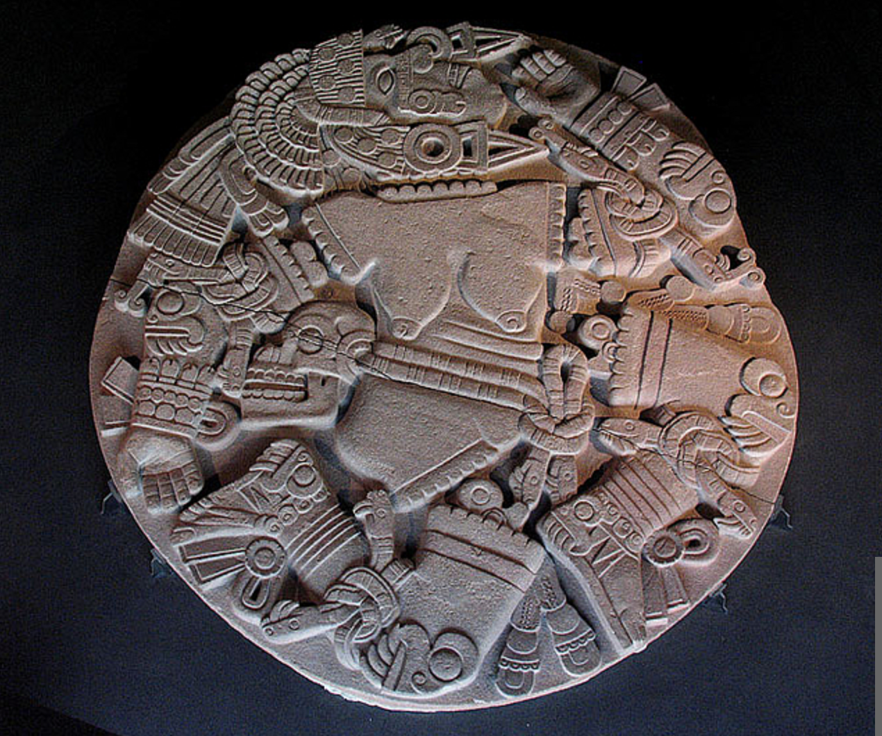 Round stone carved with intricate Mayan hieroglyphs and symbols, featuring geometric patterns and stylized animal motifs.