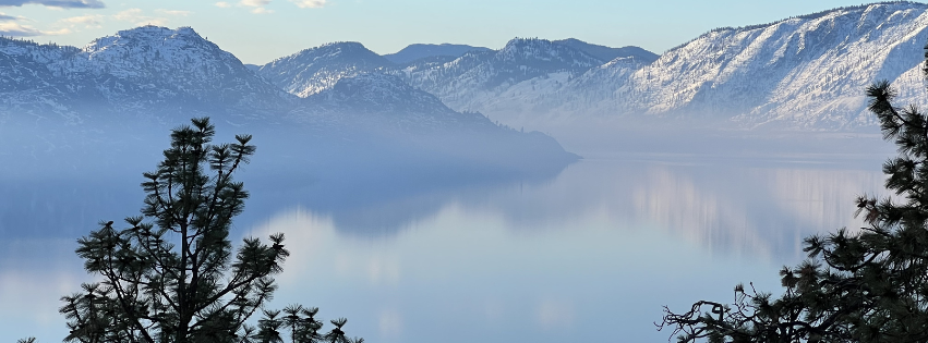 A calm lake surrounded by snow-capped mountains with pine trees in the foreground.