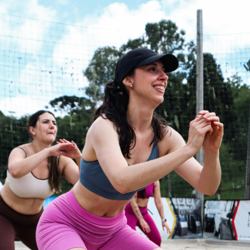 Two women exercising outdoors near a fence, with trees in the background. One woman is smiling and wearing a black cap, blue sports bra, and pink leggings. The other woman is in a beige top.