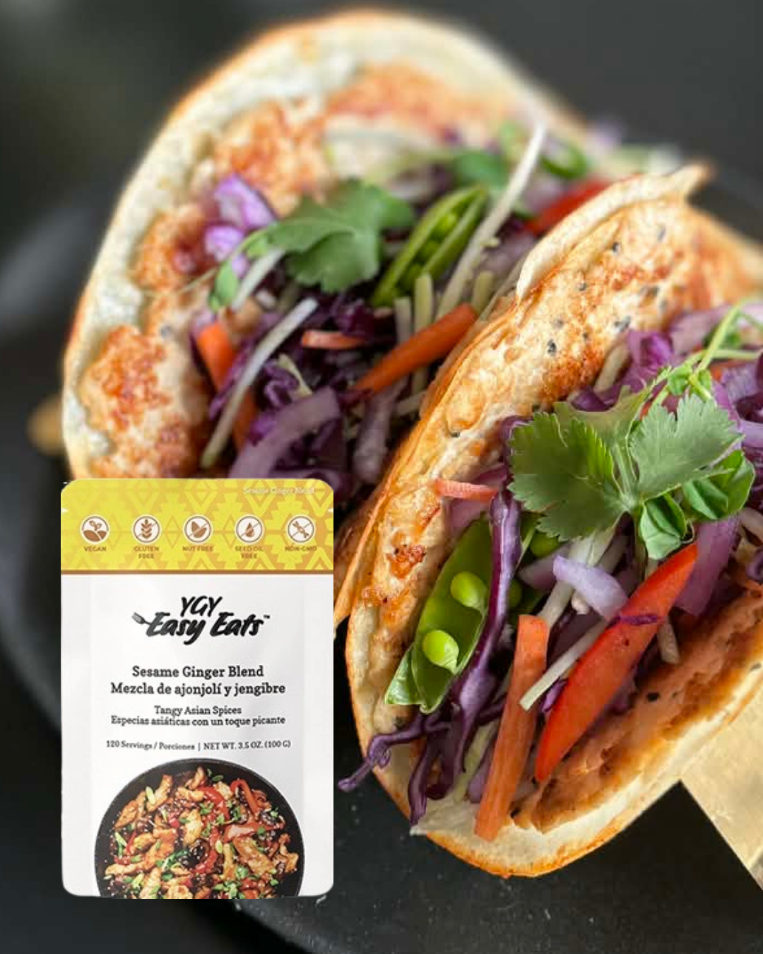 Close-up of two tacos filled with colorful vegetables such as shredded purple cabbage, orange carrots, green beans, cilantro, and other vegetables, on a black surface with a package of YCY Easy Eats Sesame Ginger Blend seasoning.