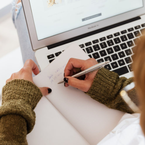 Person wearing a brown sweater writing in a notebook next to a MacBook laptop.