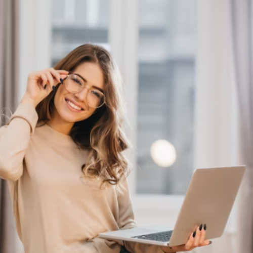 A woman smiling and adjusting her glasses while holding a laptop in her lap.