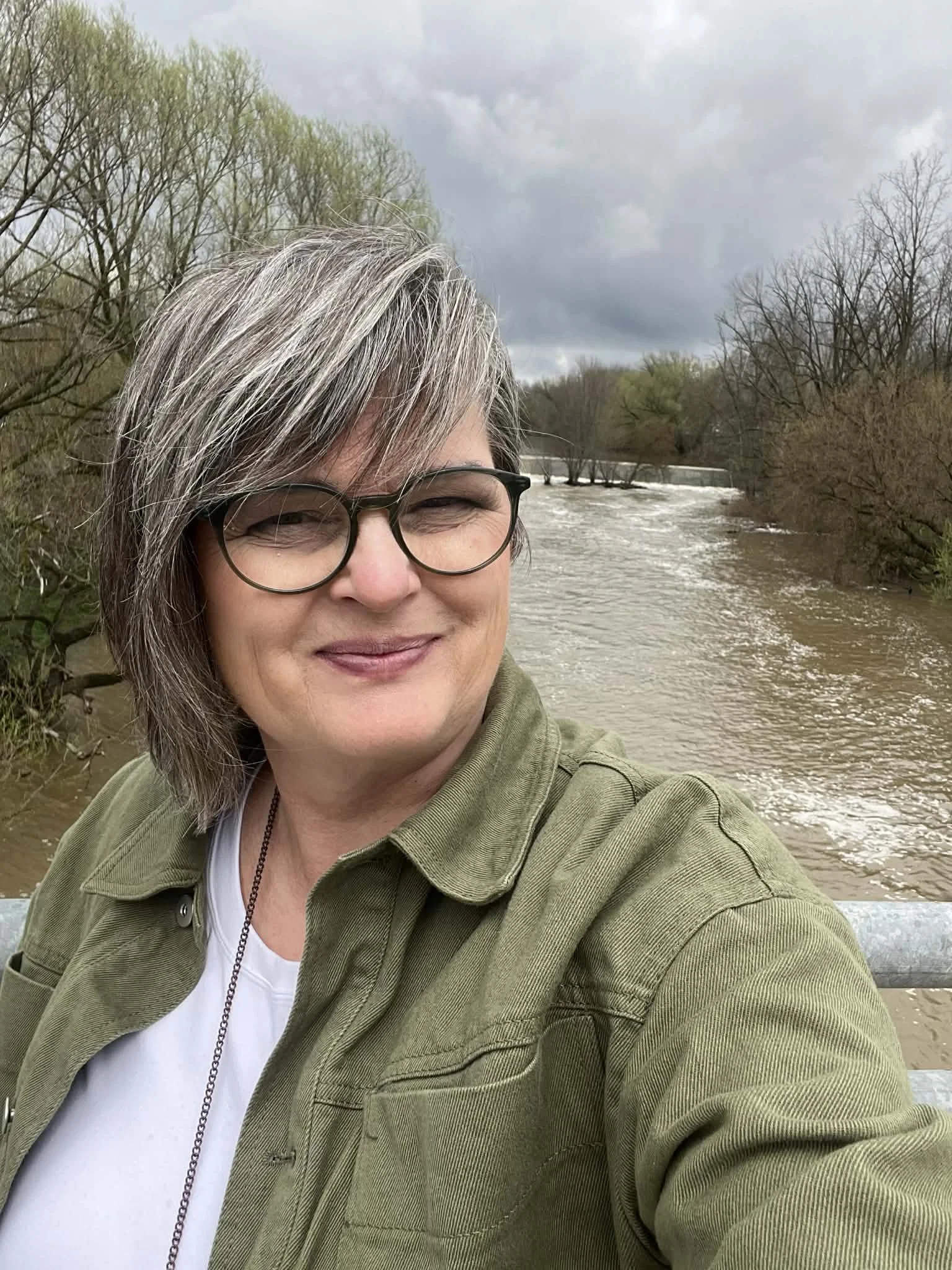 A woman with gray hair, glasses, and a green jacket taking a selfie outdoors near a river with trees and cloudy sky in the background.