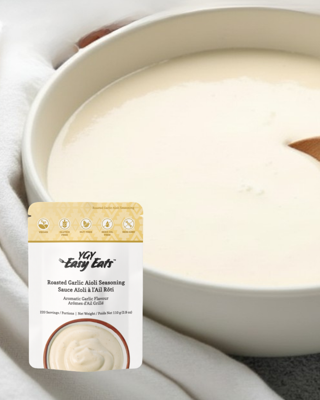 A bowl of creamy, white roasted garlic aioli with a wooden spoon partially submerged. In the foreground, there is a packet of Yu Easy Eats Roasted Garlic Aioli Seasoning.