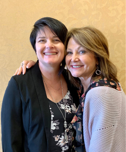 Two women smiling and hugging in front of a beige patterned wall, one wearing a black blazer and the other in a light sweater with floral details.